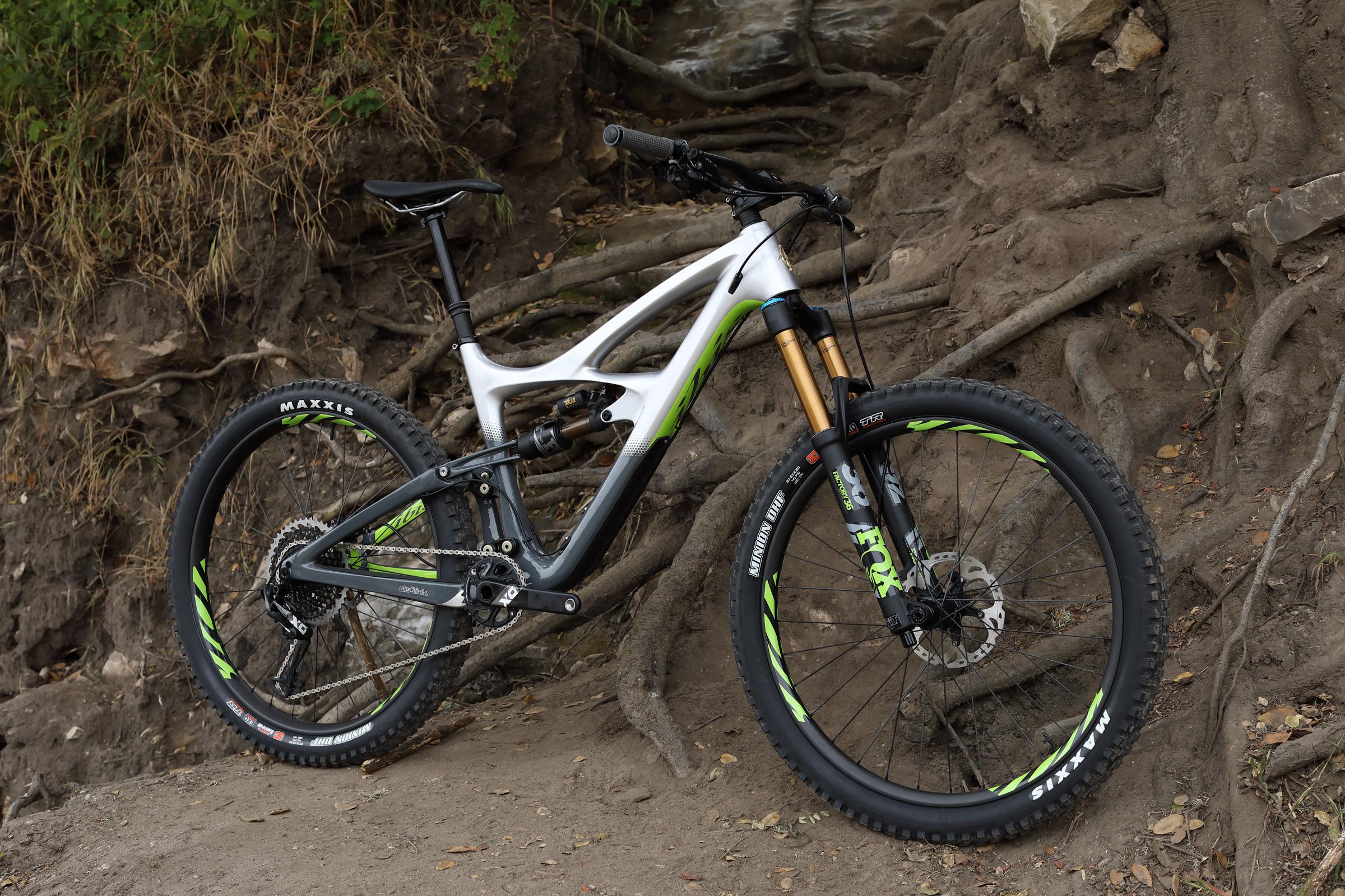 Ibis Mojo HD4: A mountain bike with a sleek white and black frame, featuring green accents, stands upright on a dirt path surrounded by tree roots. The bike is equipped with high-performance components, including a front suspension fork with gold stanchions and Maxxis tires. The background includes rocky terrain and vegetation, indicating an outdoor setting suitable for cycling.