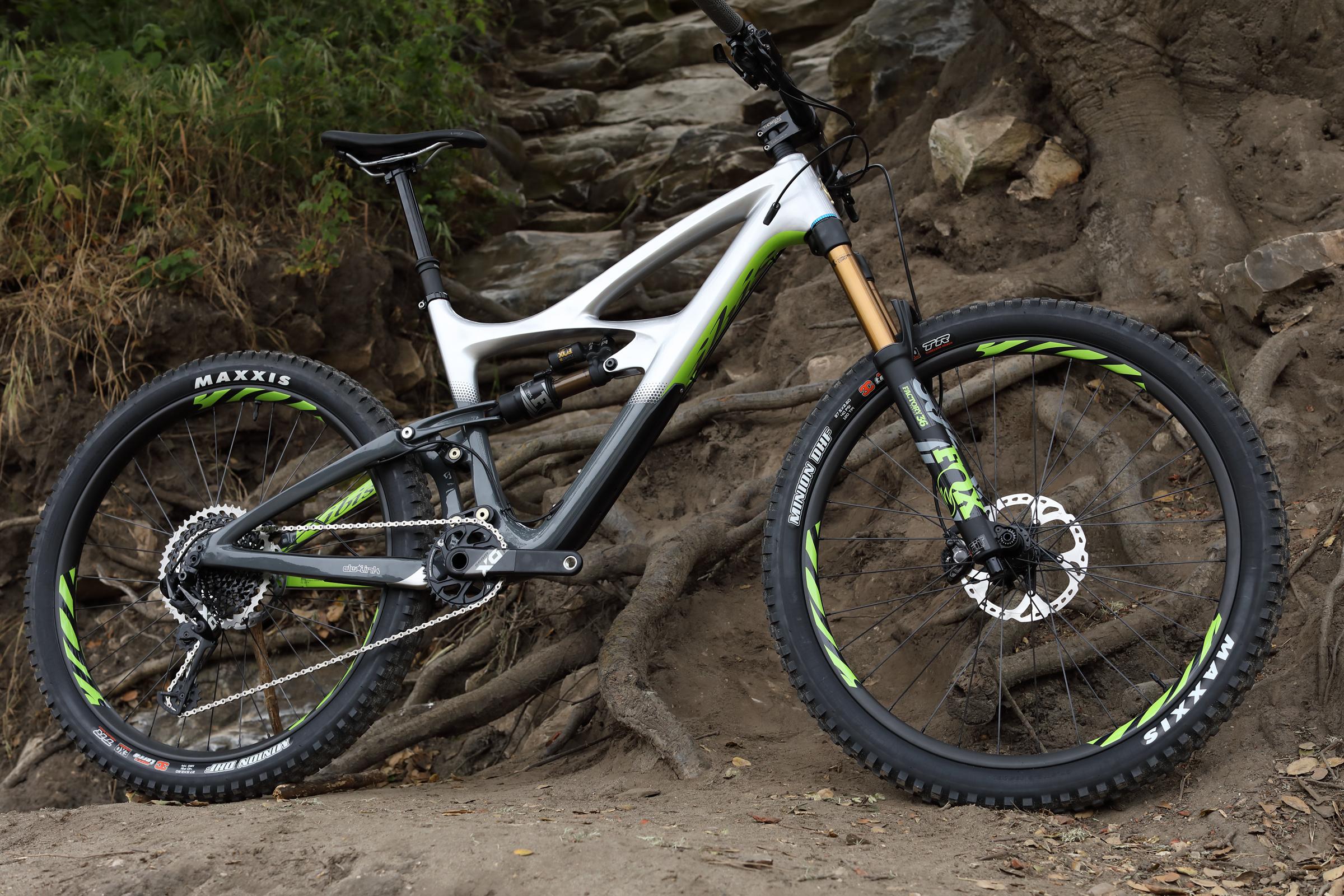 Ibis Mojo HD4: A modern mountain bike positioned on a rugged terrain, featuring a sleek white and gray frame with green accents. The bike is equipped with high-performance Maxxis tires and a front suspension fork. Surrounding the bike are natural elements like exposed roots and rocky ground, suggesting an outdoor cycling adventure.