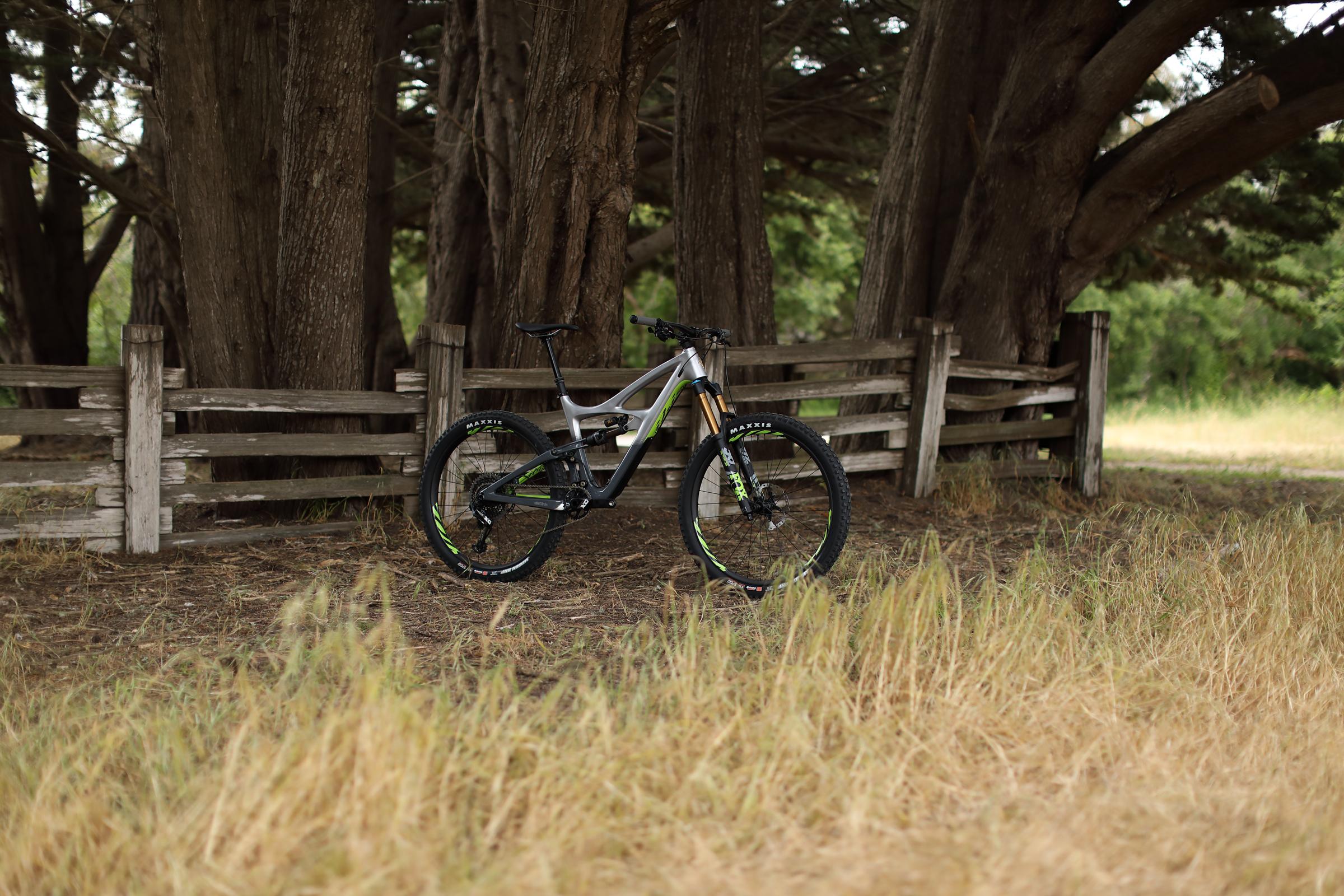 Ibis Mojo HD4: A mountain bike leaning against a wooden fence, surrounded by tall trees and golden grass. The bike features a sleek design with a silver and black frame, and is equipped with knobby tires suitable for off-road terrain.