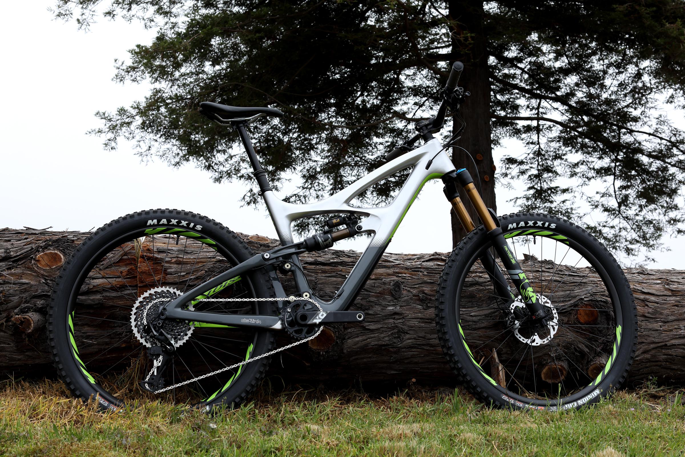 Ibis Mojo HD4: Mountain bike leaning against a large log, set in a grassy field with trees in the background. The bike features a sleek design with a white and black frame, green accents on the wheels, and sturdy tires suitable for off-road terrain.