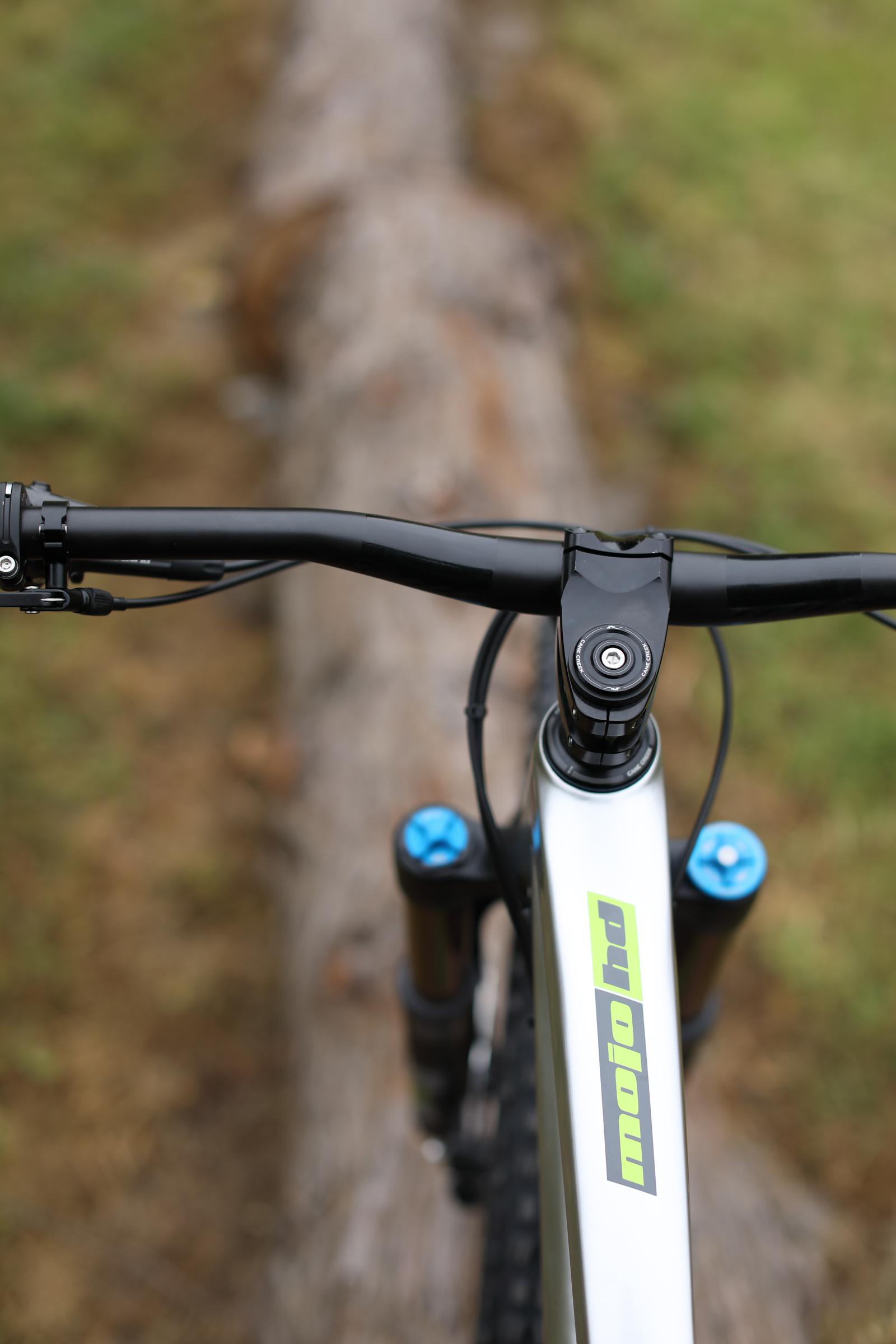 Ibis Mojo HD4: Close-up view of a mountain bike's handlebars and frame, showcasing the handlebar grips, stem, and logo. The bike is positioned on a log in a grassy outdoor setting, suggesting an adventure in nature.