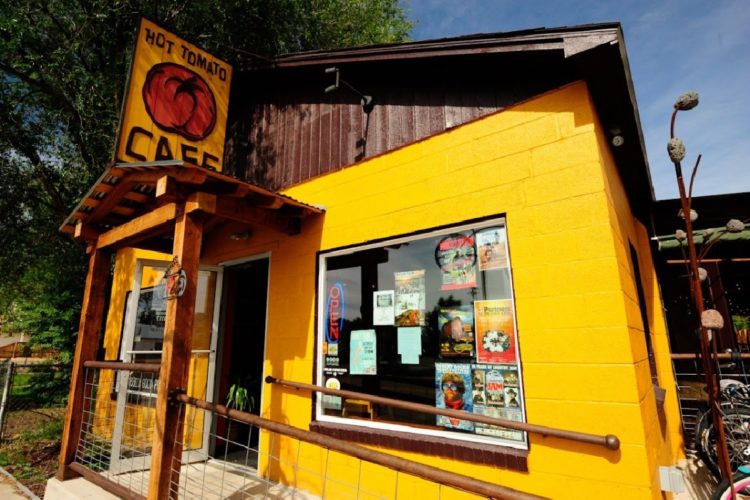 A brightly colored yellow cafe building with a wooden entrance and a sign that reads "Hot Tomato Cafe." The window displays various posters and notices, and an "Open" sign is illuminated. Lush trees are visible in the background, suggesting a lively outdoor setting.