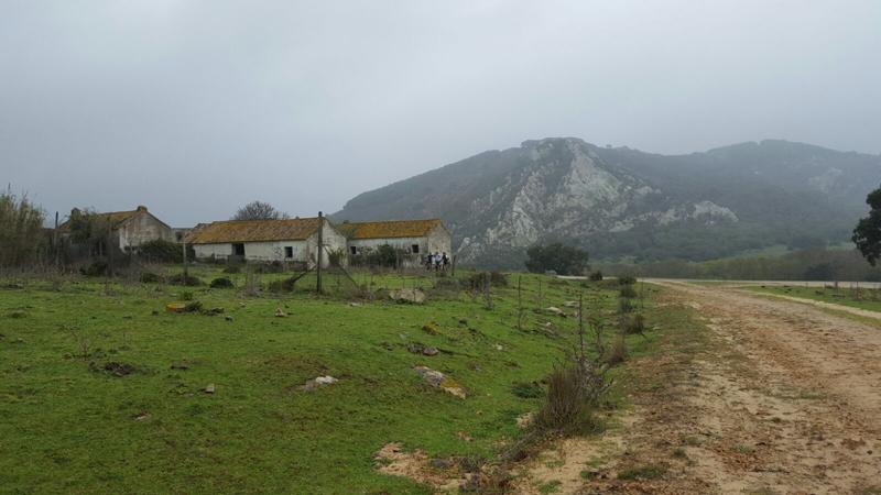 A rural landscape featuring an old, weathered building with a mossy, brown roof, set against a backdrop of a cloudy sky and a mountainous terrain. The foreground includes a dirt road leading to the structure, surrounded by grassy fields and scattered rocks.
