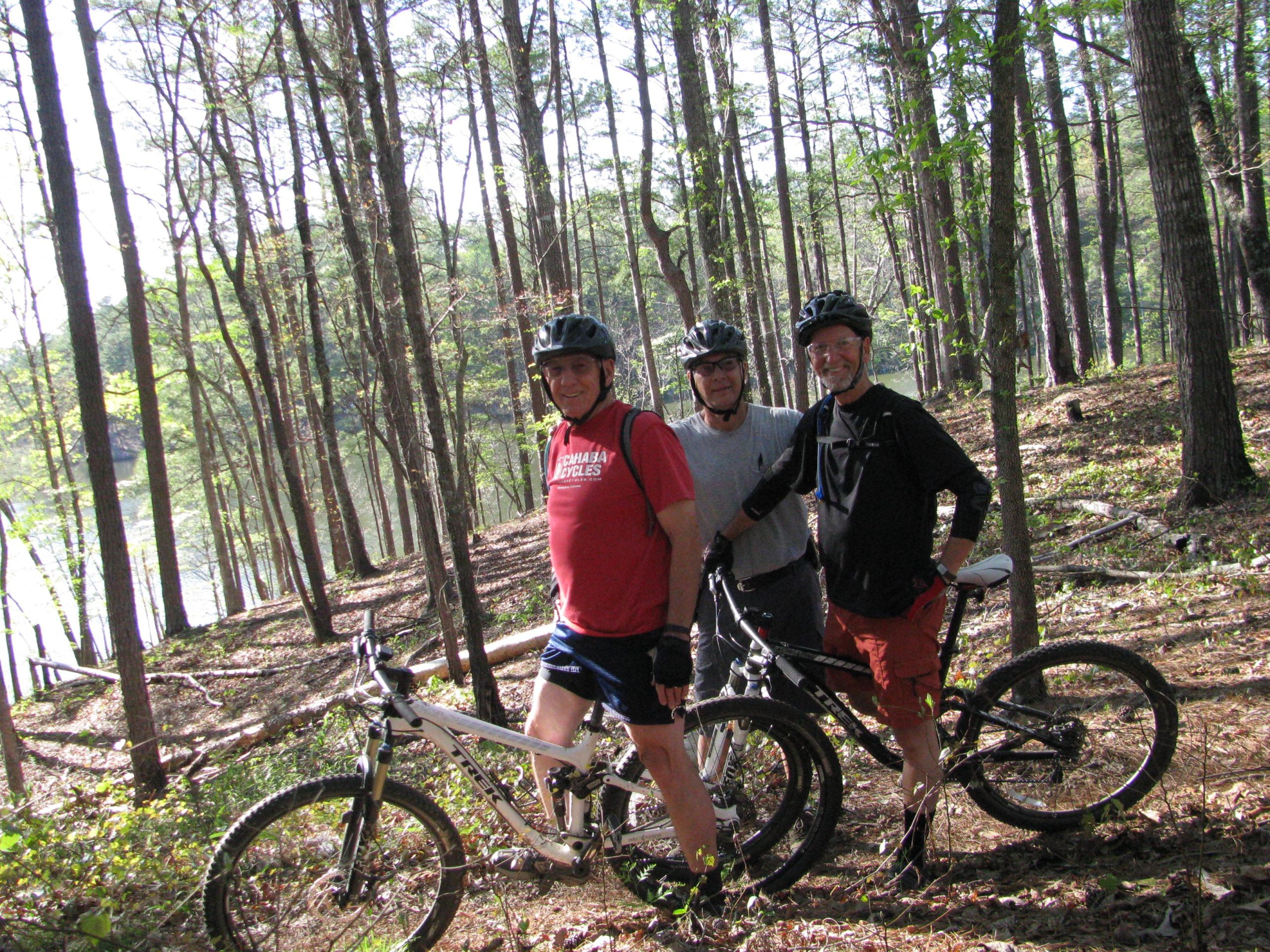 Three mountain bikers pose for a photo in a wooded area near a body of water. They are wearing helmets and cycling gear. The scene is bright and sunny, with tall trees surrounding them. Sylaward mountain bike trail.
