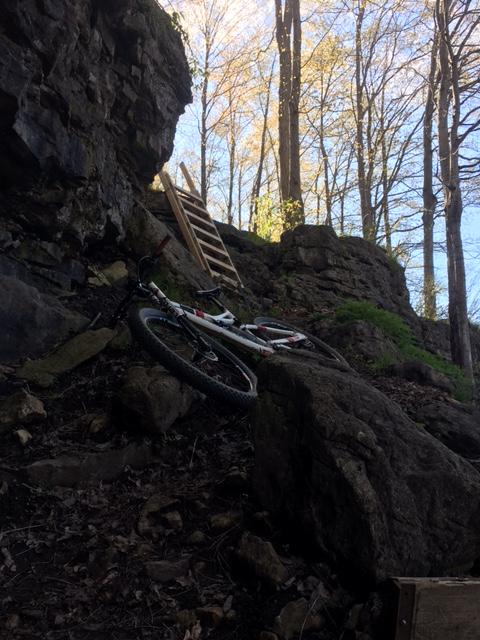A mountain bike rests on rocky, uneven terrain with large boulders and a wooden ladder visible in the background, surrounded by trees in a wooded area under a clear sky. Inglis falls West rock mountain bike trail.