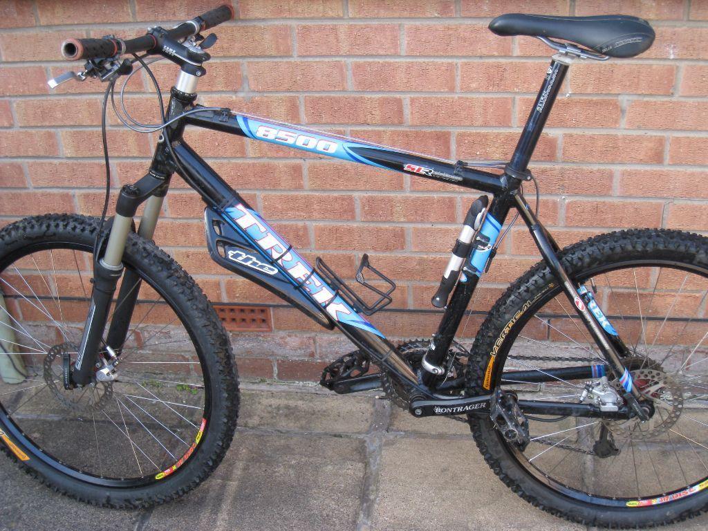 Trek 8500 LT: A black and blue mountain bike leaning against a brick wall. The bike features wide, knobby tires, a front suspension fork, and a sleek frame design. The handlebars have rubber grips, and there is a water bottle holder attached to the frame. The area in the background includes a portion of a brick wall and a paved surface.
