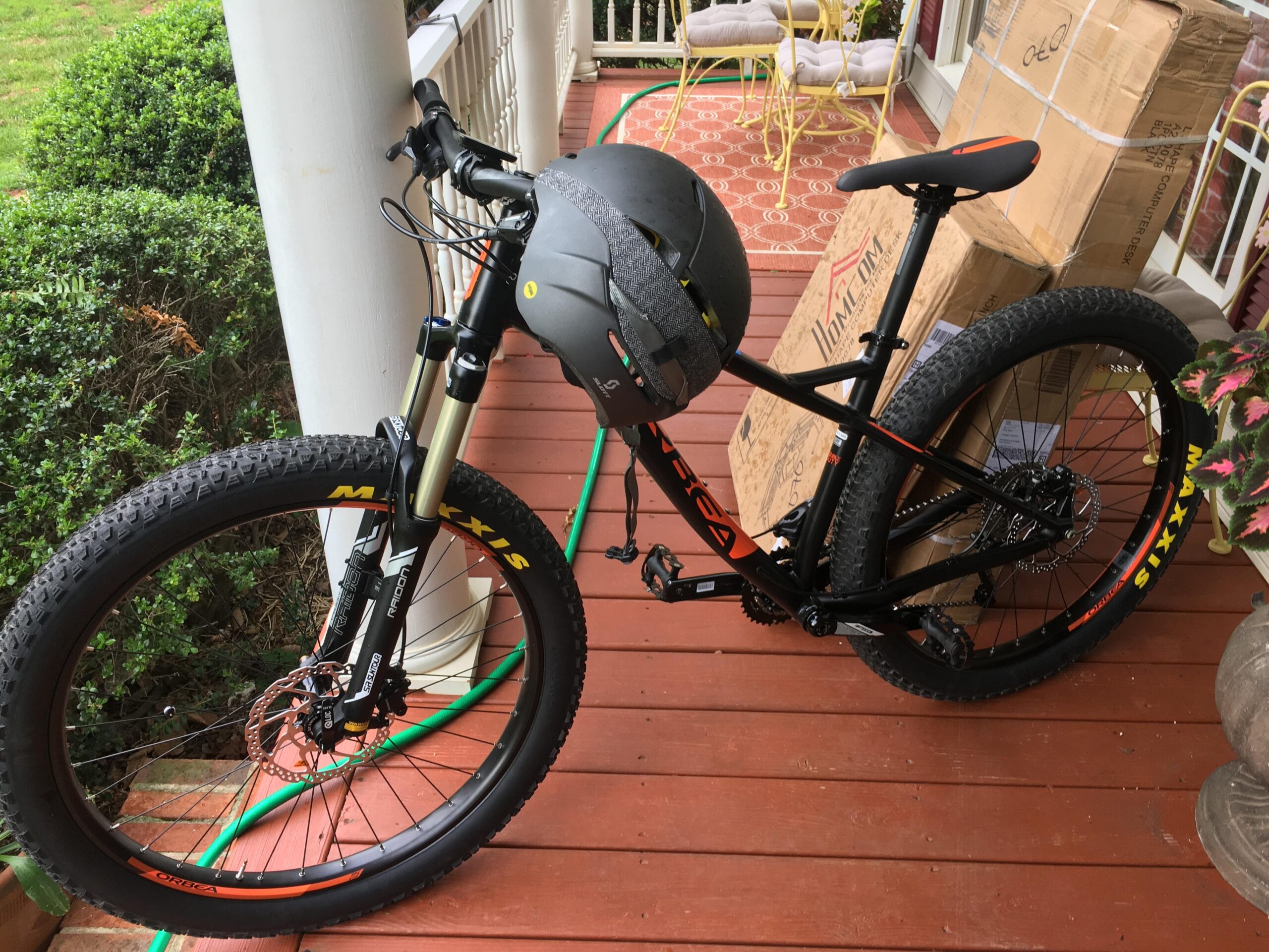 Orbea Loki: A mountain bike leaning against a porch railing, equipped with a gray helmet on the handlebars. The bike features thick tires and disc brakes. In the background, there are boxes stacked on the porch and green bushes visible.