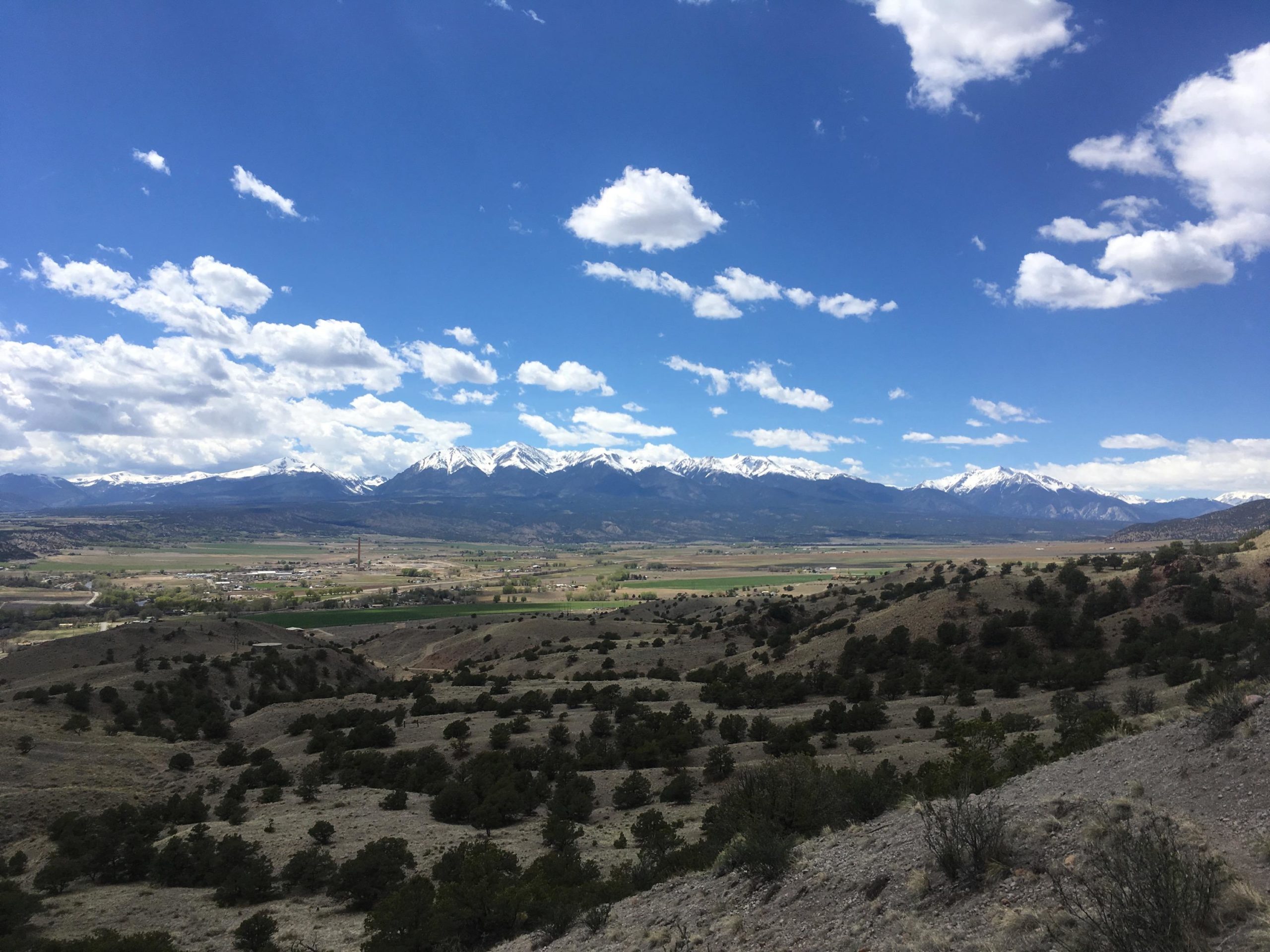 A panoramic view of a mountainous landscape under a bright blue sky with scattered clouds. Snow-capped peaks rise majestically in the background, while rolling hills and patches of greenery fill the foreground. The scene captures a serene valley with hints of agriculture, indicative of a rural area surrounded by natural beauty. Arkansas Hills mountain bike trail.