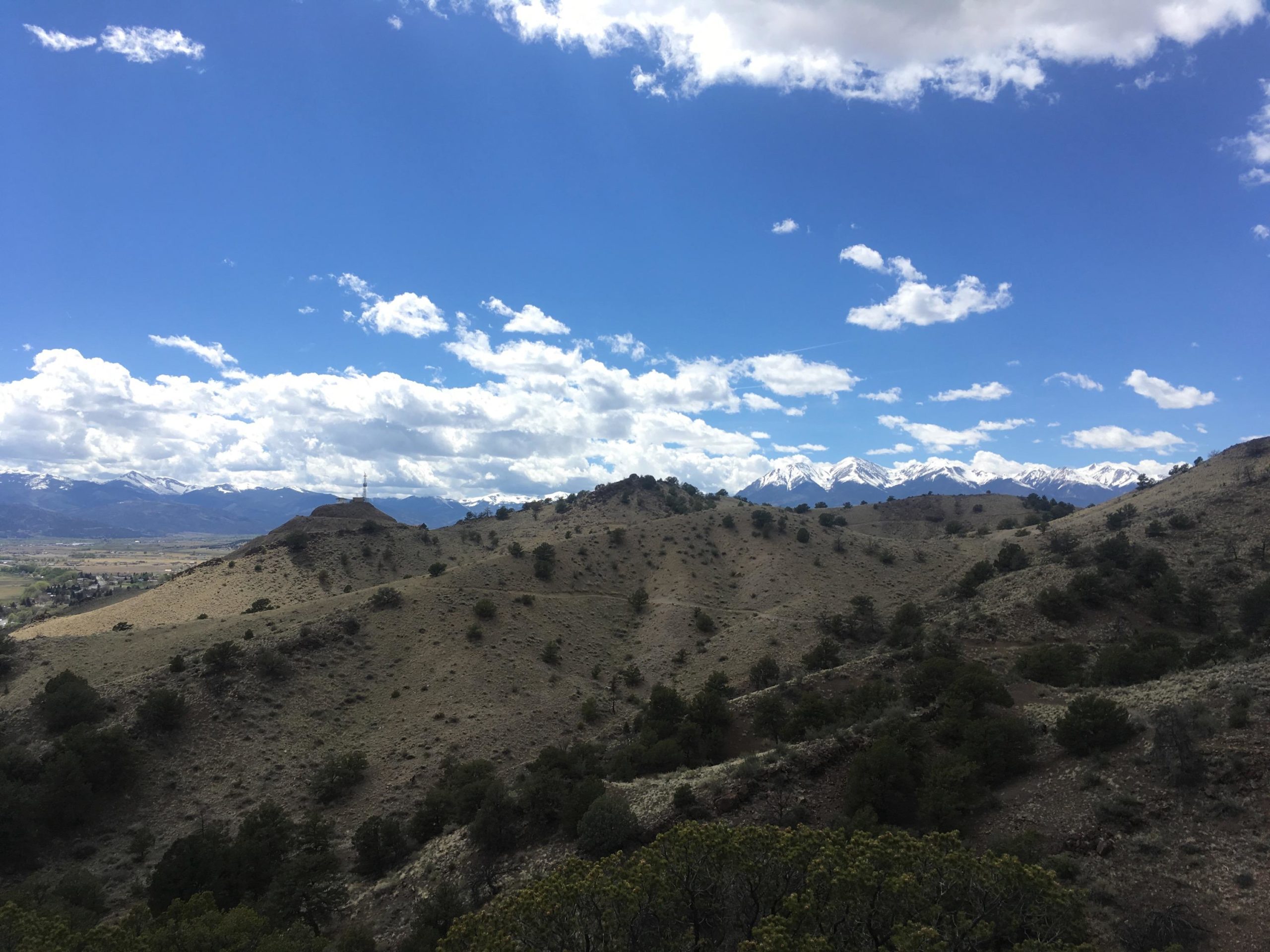 A scenic landscape view featuring rolling hills and sparse vegetation under a bright blue sky with fluffy white clouds. In the background, snow-capped mountains rise majestically against the horizon, creating a dramatic contrast with the more arid foreground. Arkansas Hills mountain bike trail.