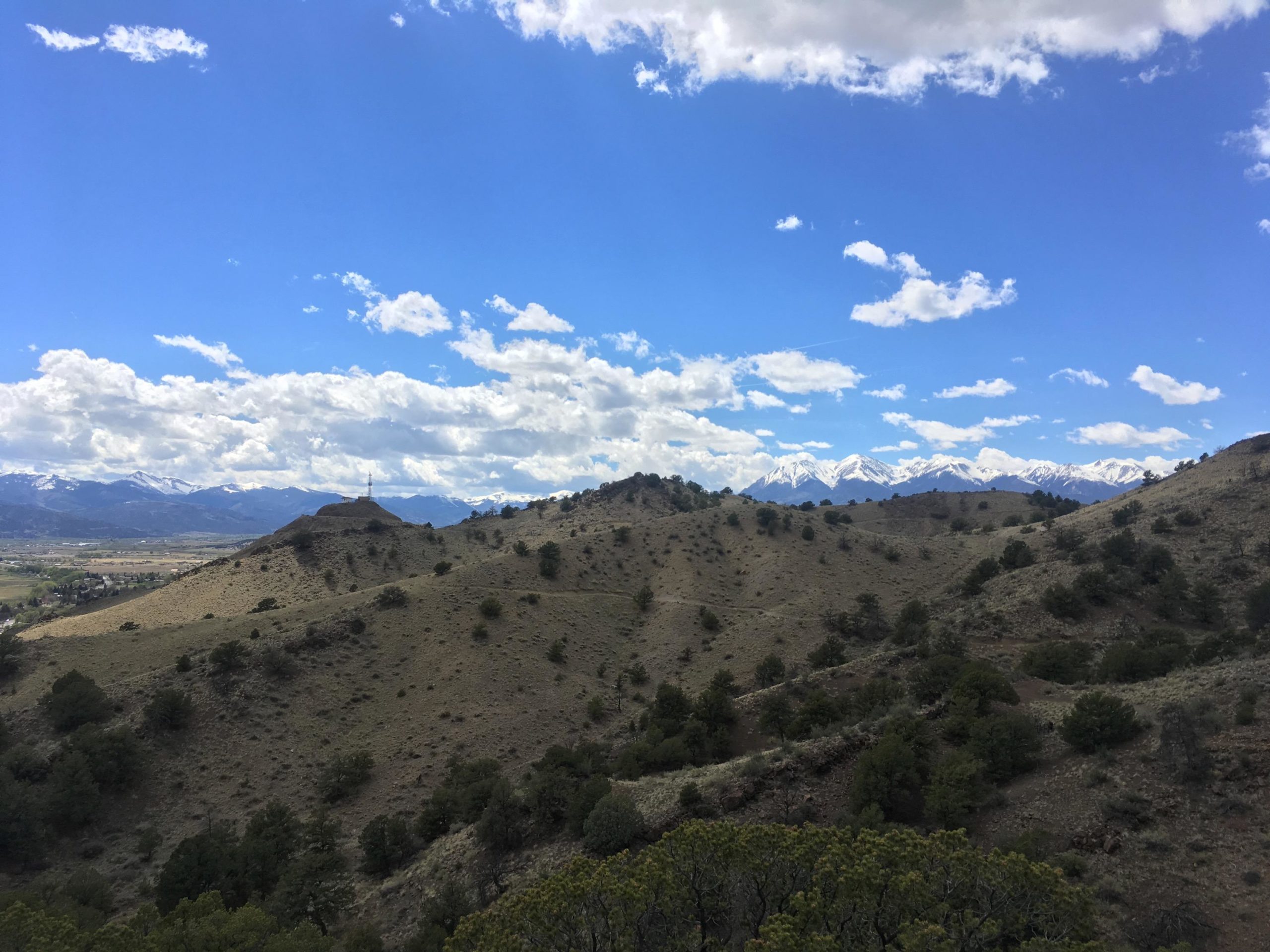 A landscape view featuring rolling hills with sparse vegetation, set against a backdrop of snow-capped mountains under a blue sky dotted with clouds. A communication tower is visible on a hill in the foreground. Arkansas Hills mountain bike trail.