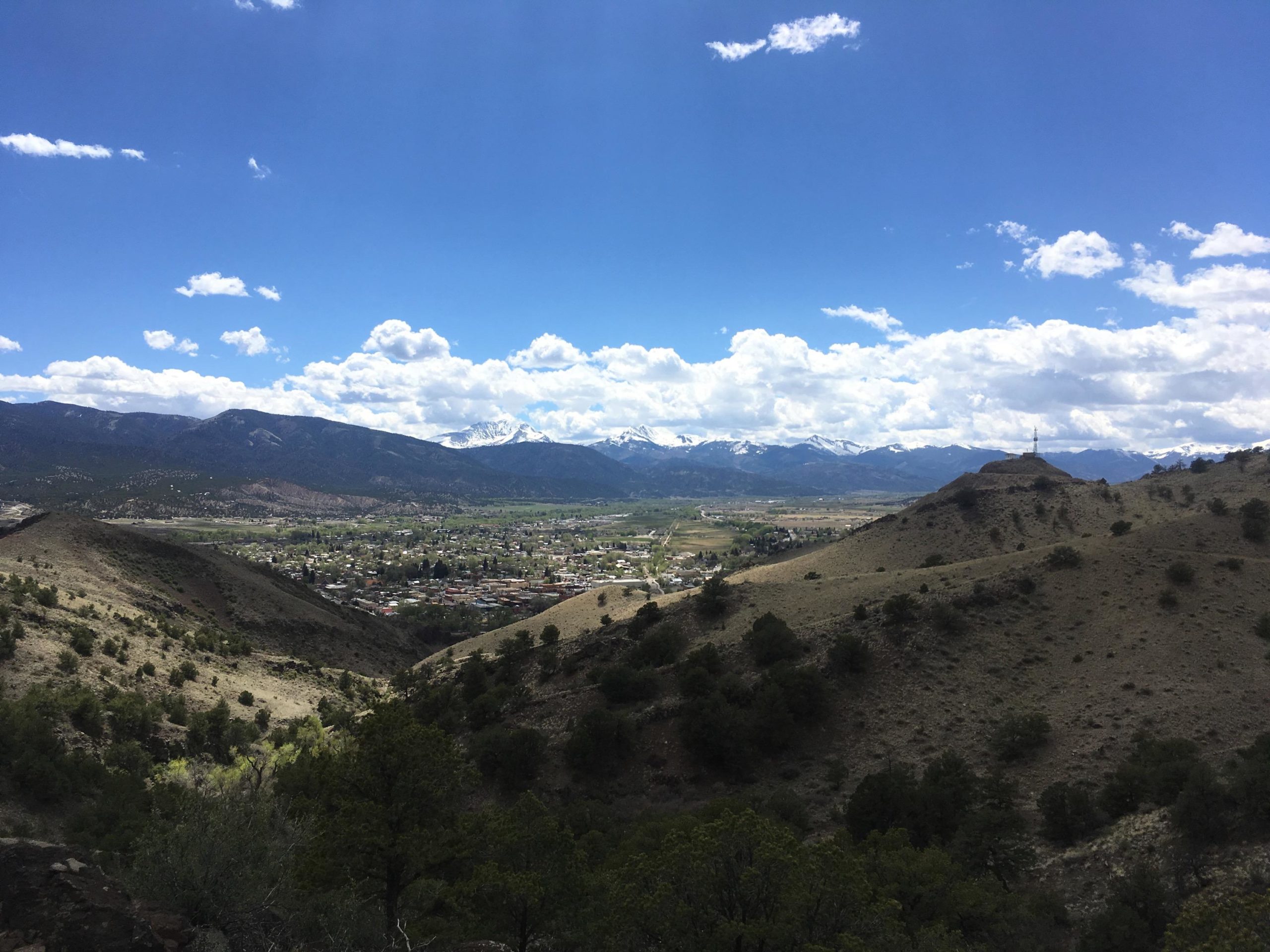 A panoramic view of a valley surrounded by rolling hills and mountains under a clear blue sky with scattered clouds. The foreground features gentle slopes covered in shrubs and trees, while the background showcases snow-capped peaks. A small town is visible in the valley, nestled among the landscape. Arkansas Hills mountain bike trail.