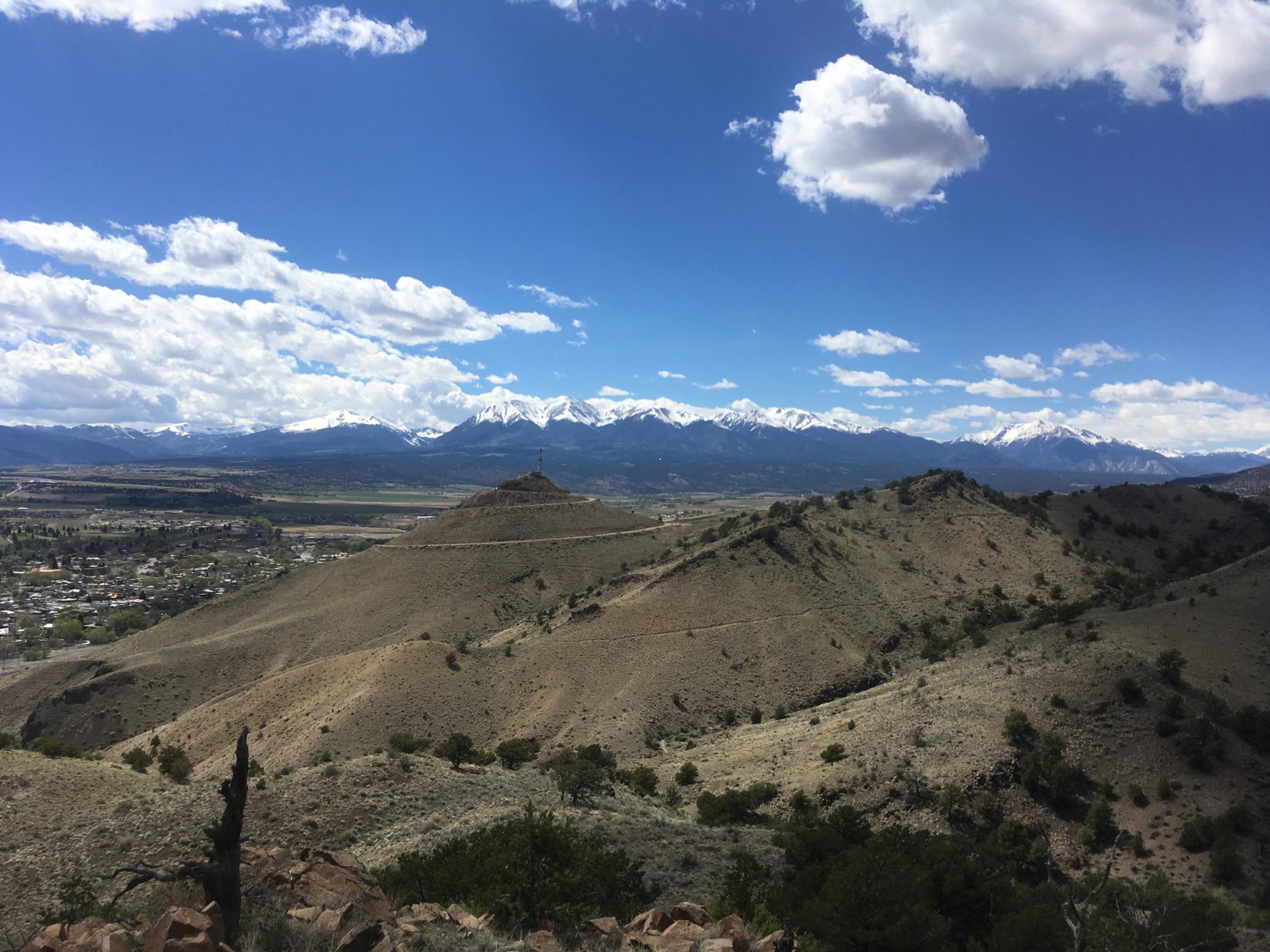A panoramic view of a mountainous landscape under a partly cloudy sky. In the foreground, rolling hills lead to a small peak with a tower at the top. In the background, dramatic snow-capped mountains rise against a vibrant blue sky. A small town is visible in the valley below, surrounded by greenery. Arkansas Hills mountain bike trail.