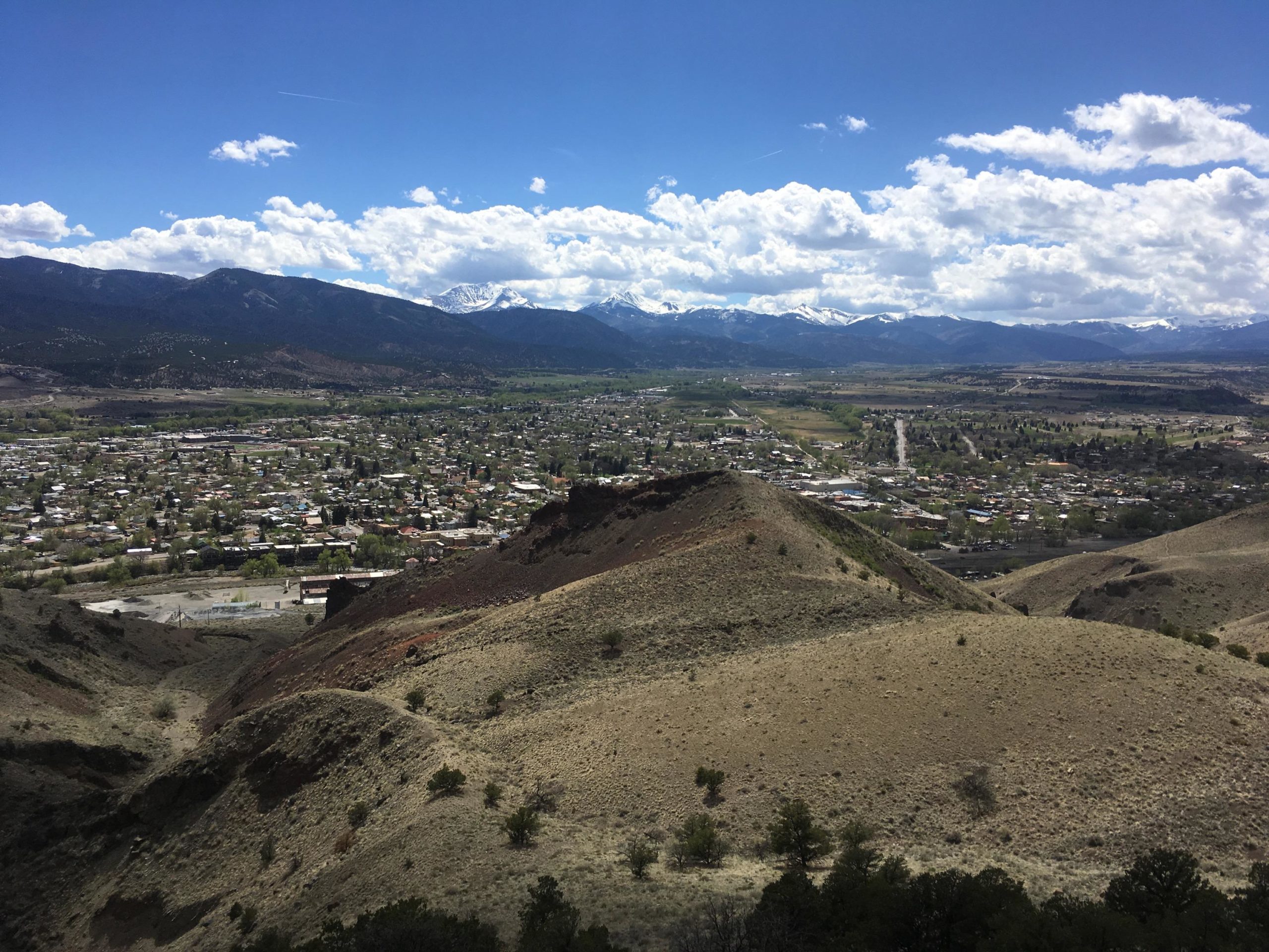 A panoramic view of a valley surrounded by mountains, featuring a small town with scattered houses and green trees. The foreground includes rolling hills with dry grass and small shrubs, while the background showcases snow-capped mountains under a partly cloudy blue sky. Arkansas Hills mountain bike trail.