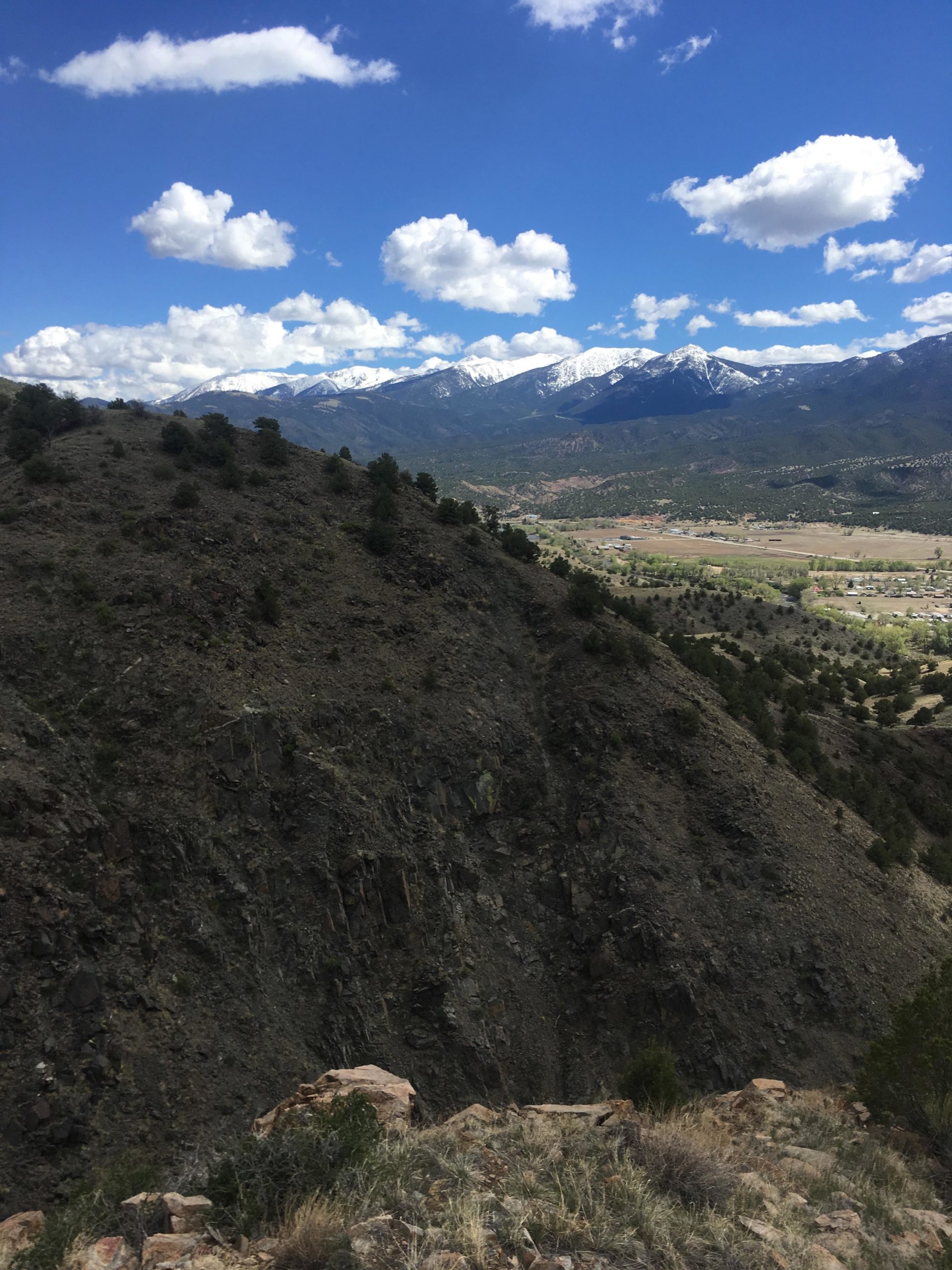 A panoramic view of a mountainous landscape featuring rugged hills in the foreground, snow-capped peaks in the background, and a clear blue sky with fluffy white clouds. The scene showcases a mix of greenery and rocky terrain, indicating a natural, unspoiled environment. Arkansas Hills mountain bike trail.