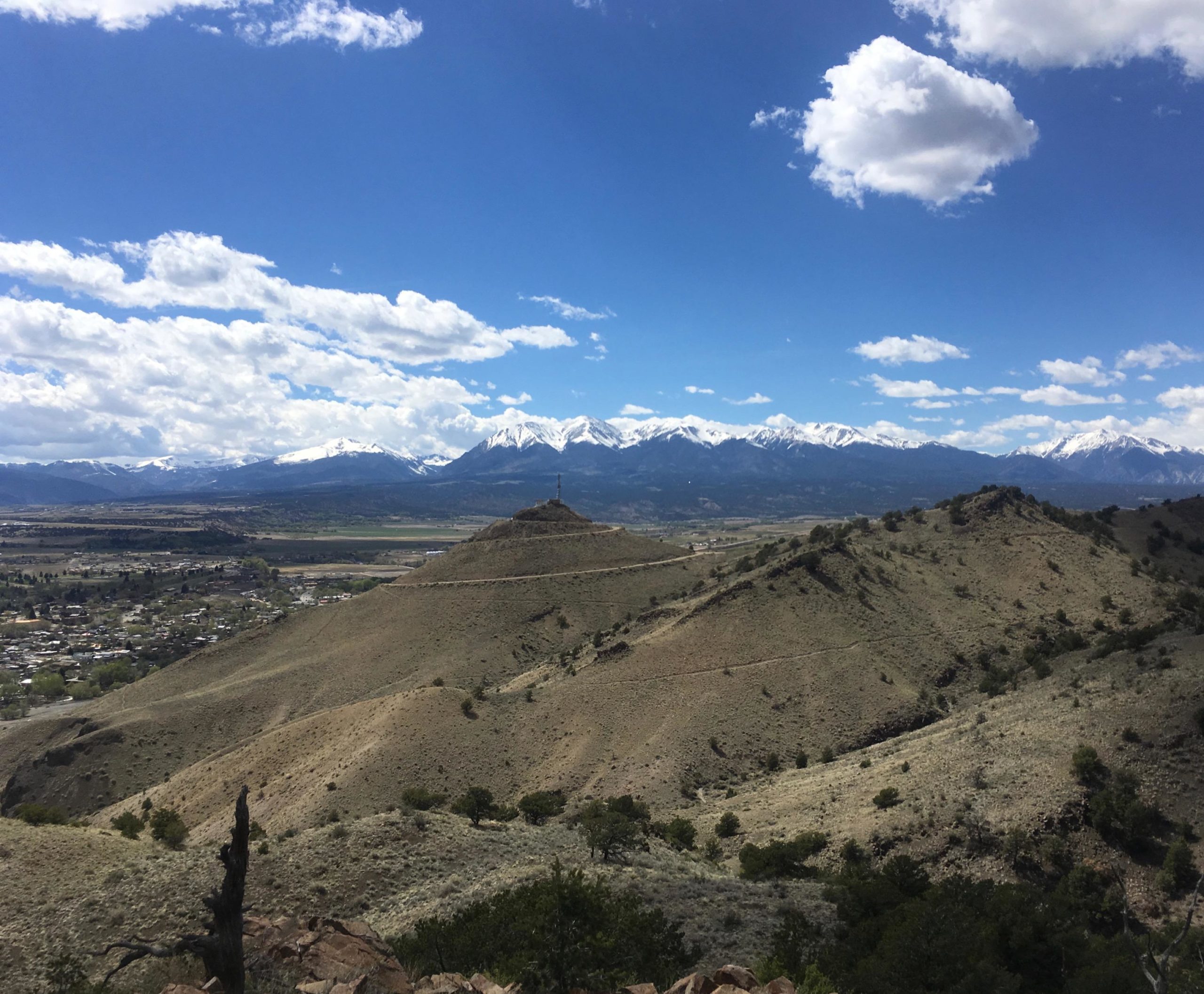 A scenic landscape showcasing rolling hills leading to a distant mountain range capped with snow. The sky is bright blue with fluffy white clouds scattered across it. In the foreground, grassy hills and sparse trees are visible, while a small town is nestled in the valley below, providing a contrast to the rugged terrain. Arkansas Hills mountain bike trail.
