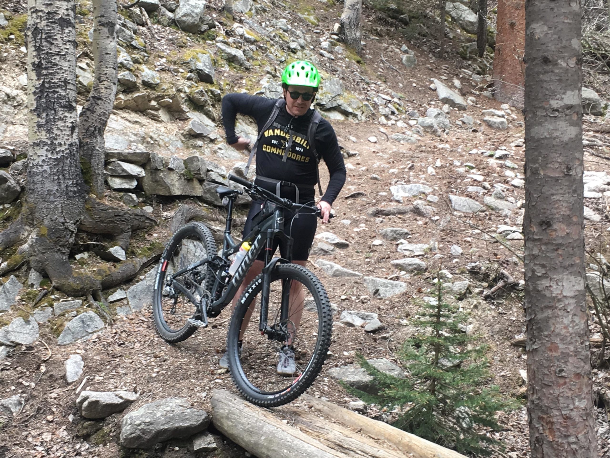 A person in a green helmet is standing next to a mountain bike on a rocky trail surrounded by trees. The individual is wearing a black shirt with yellow lettering and appears to be adjusting their gear, preparing to continue their ride. Colorado Trail: Mount Princeton to Avalanche Trailhead / Collegiate Peaks Wilderness mountain bike trail.