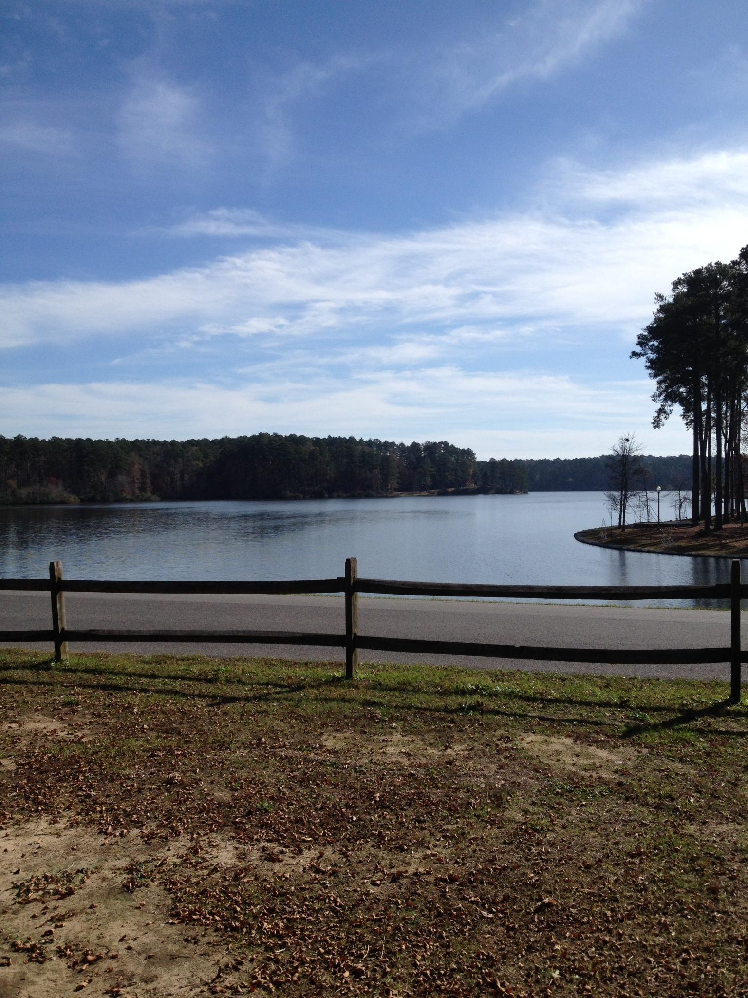 A serene view of a calm lake bordered by a wooden fence, surrounded by tall trees. The sky is partly cloudy with patches of blue, reflecting on the water's surface. Sunlight casts gentle shadows on the grassy bank in the foreground, which is strewn with fallen leaves. Bonita Lakes mountain bike trail.