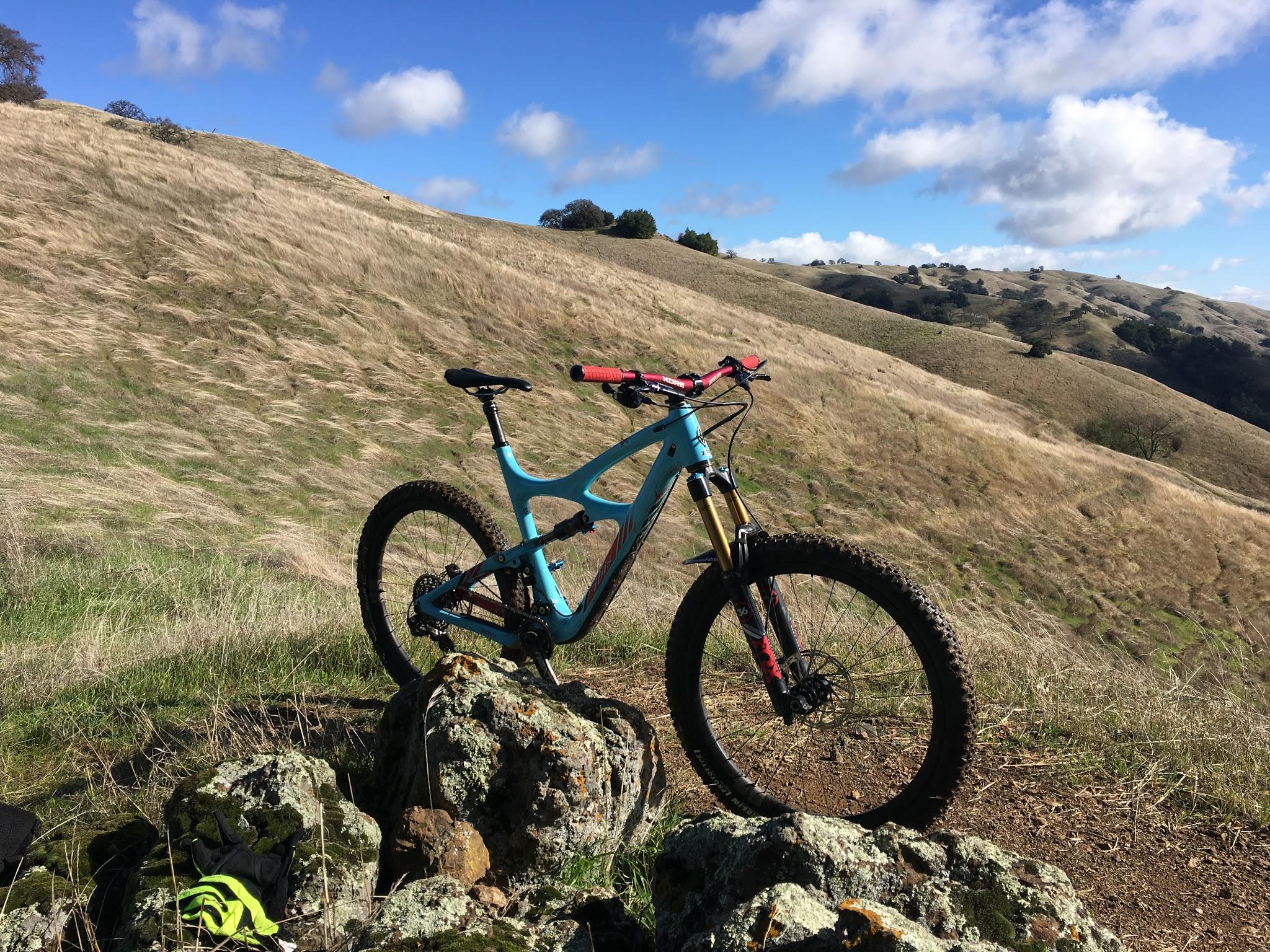 Ibis Mojo HD3: A mountain bike leaning on a rock in a grassy landscape with rolling hills under a blue sky dotted with clouds.