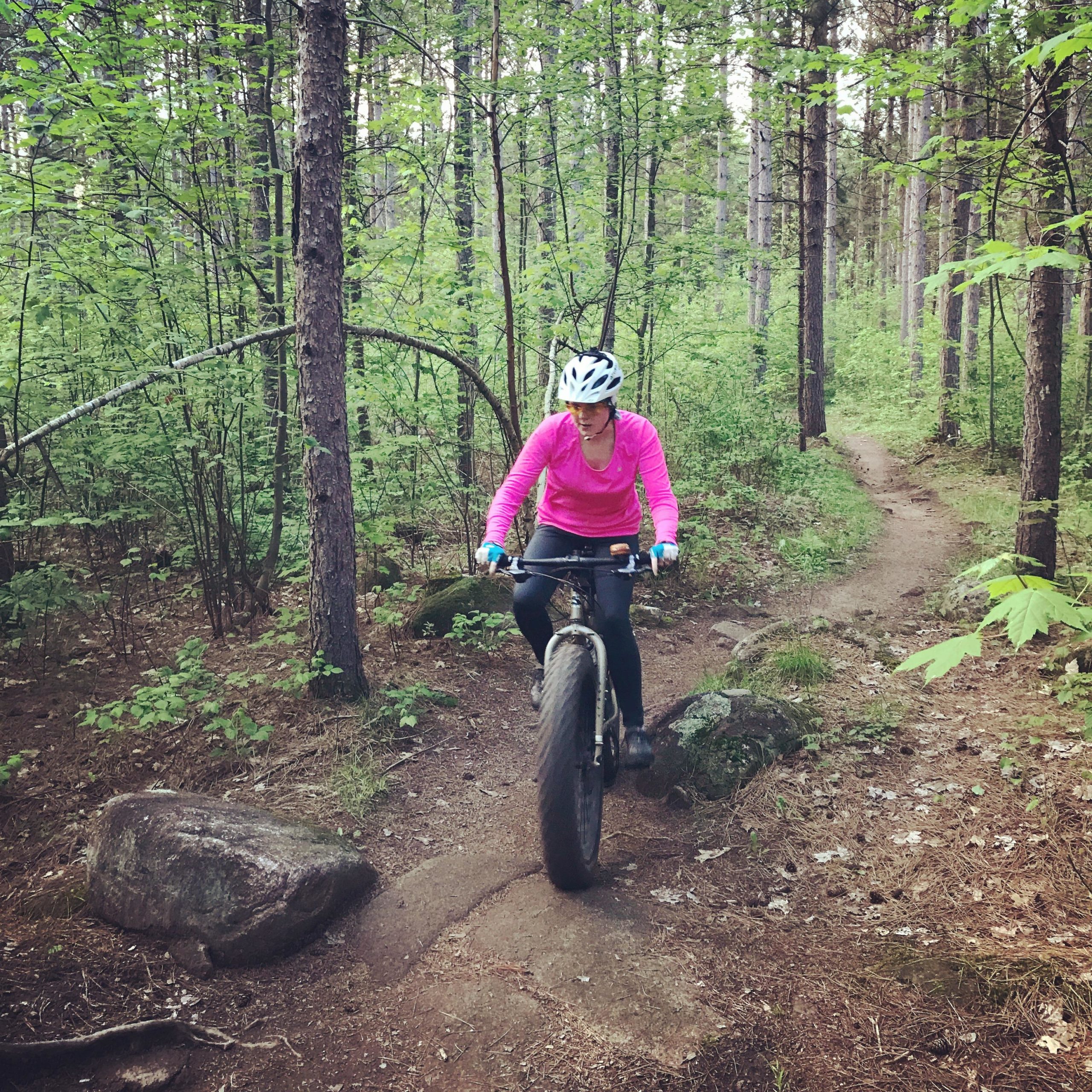 A person wearing a pink long-sleeve shirt and a helmet rides a fat bike along a narrow trail in a lush forest. The surroundings include trees, green foliage, and a rocky path, suggesting an outdoor adventure in nature. Nine Mile mountain bike trail.