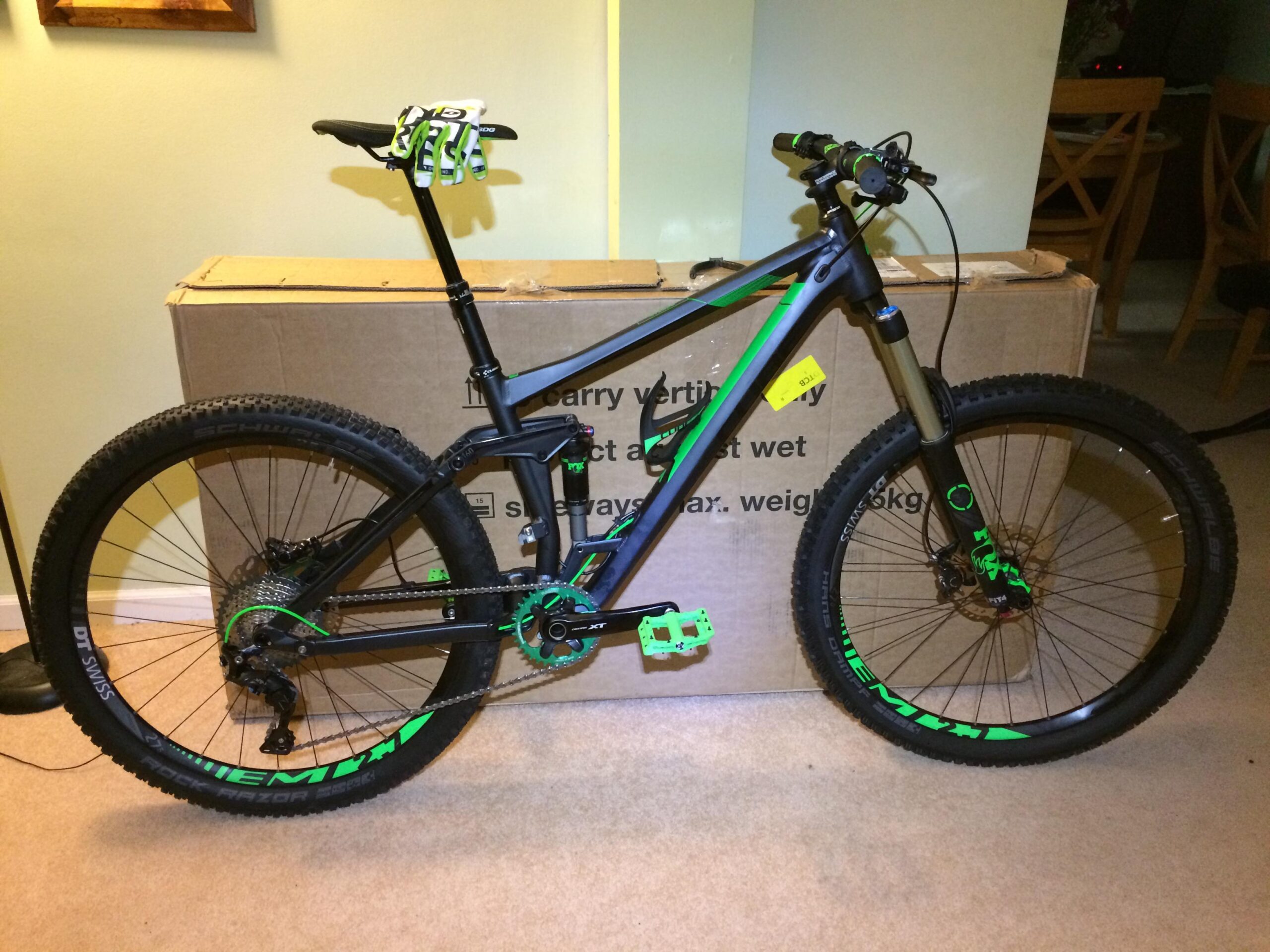 Cube Cube Stereo 160 HPA SL: A black and green mountain bike is leaning against a cardboard box. The bike features thick tires, a suspension fork, and green accents on the pedals, handlebars, and frame. A pair of cycling gloves is resting on the bike's seat. The background includes a softly lit room with wooden furniture.