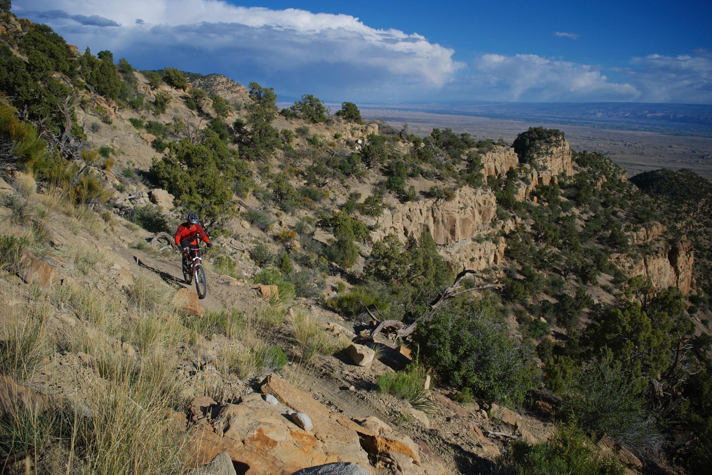 A mountain biker riding along a rugged trail on a hillside, surrounded by rocky terrain and sparse vegetation. In the background, a vast landscape stretches out under a blue sky with white clouds. Sarlacc mountain bike trail.