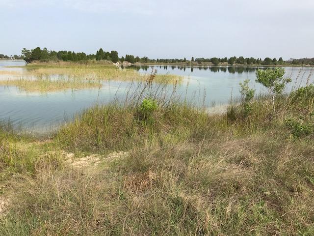 A tranquil landscape featuring a calm body of water with grassy banks, surrounded by sparse vegetation and small islands in the distance. The sky is partly cloudy, creating a peaceful outdoor scene. Open Quarry Rd. mountain bike trail.
