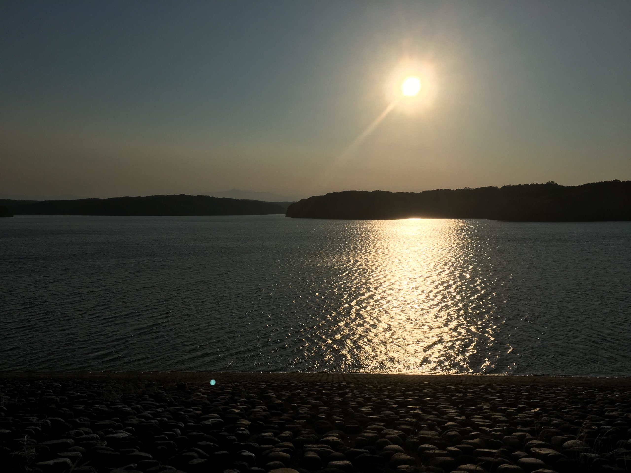 A serene sunset over a calm body of water, with the sun illuminating the surface and creating a shimmering reflection. Dark silhouettes of trees line the horizon, and a rocky shoreline is visible in the foreground. Sayama/Tama Lake mountain bike trail.