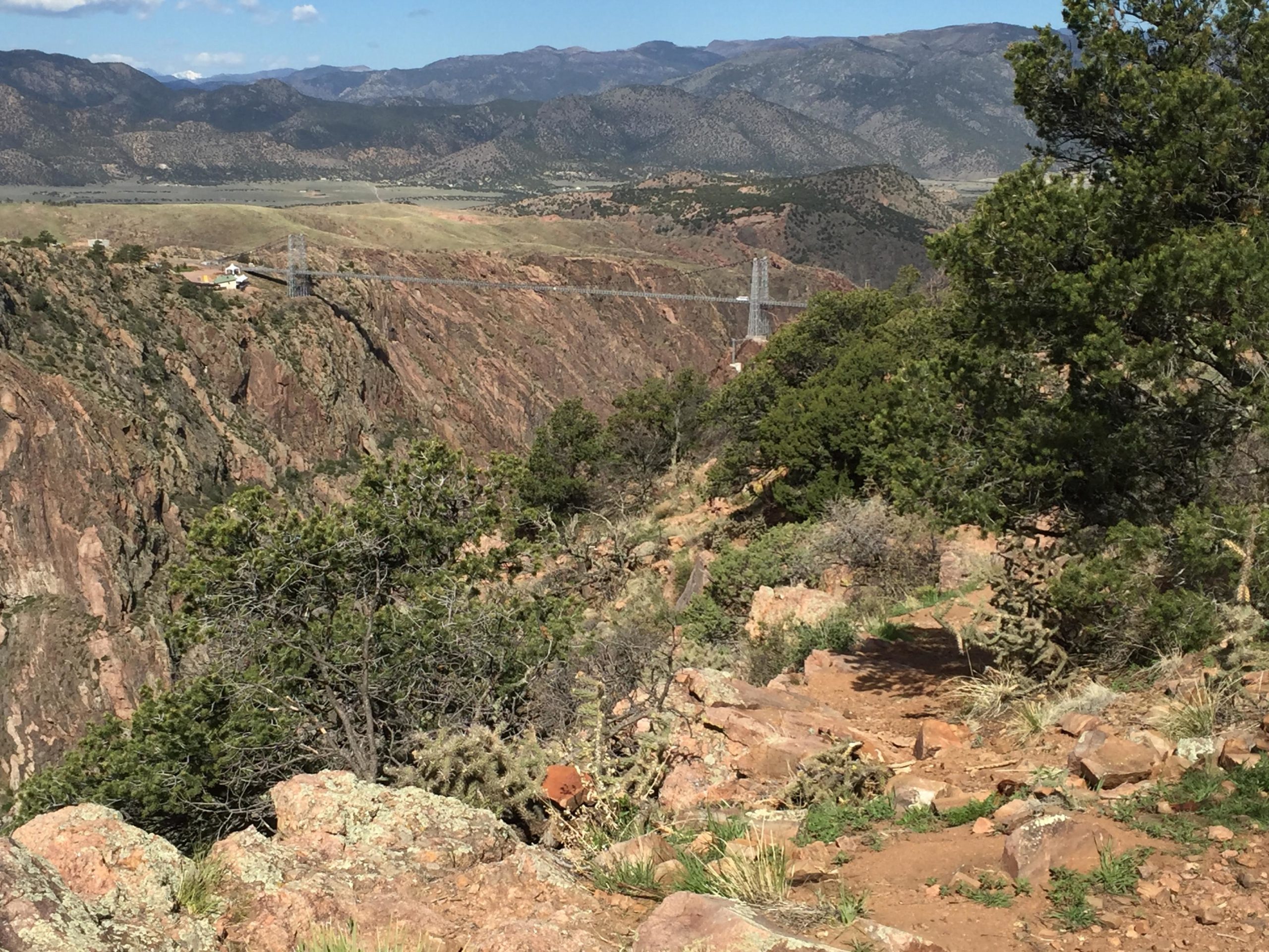 A scenic view of a mountainous landscape featuring a suspension bridge spanning a deep canyon. The foreground includes rocky terrain and sparse vegetation, while in the background, rolling mountains and a clear blue sky can be seen. Royal Gorge Park Trail System mountain bike trail.