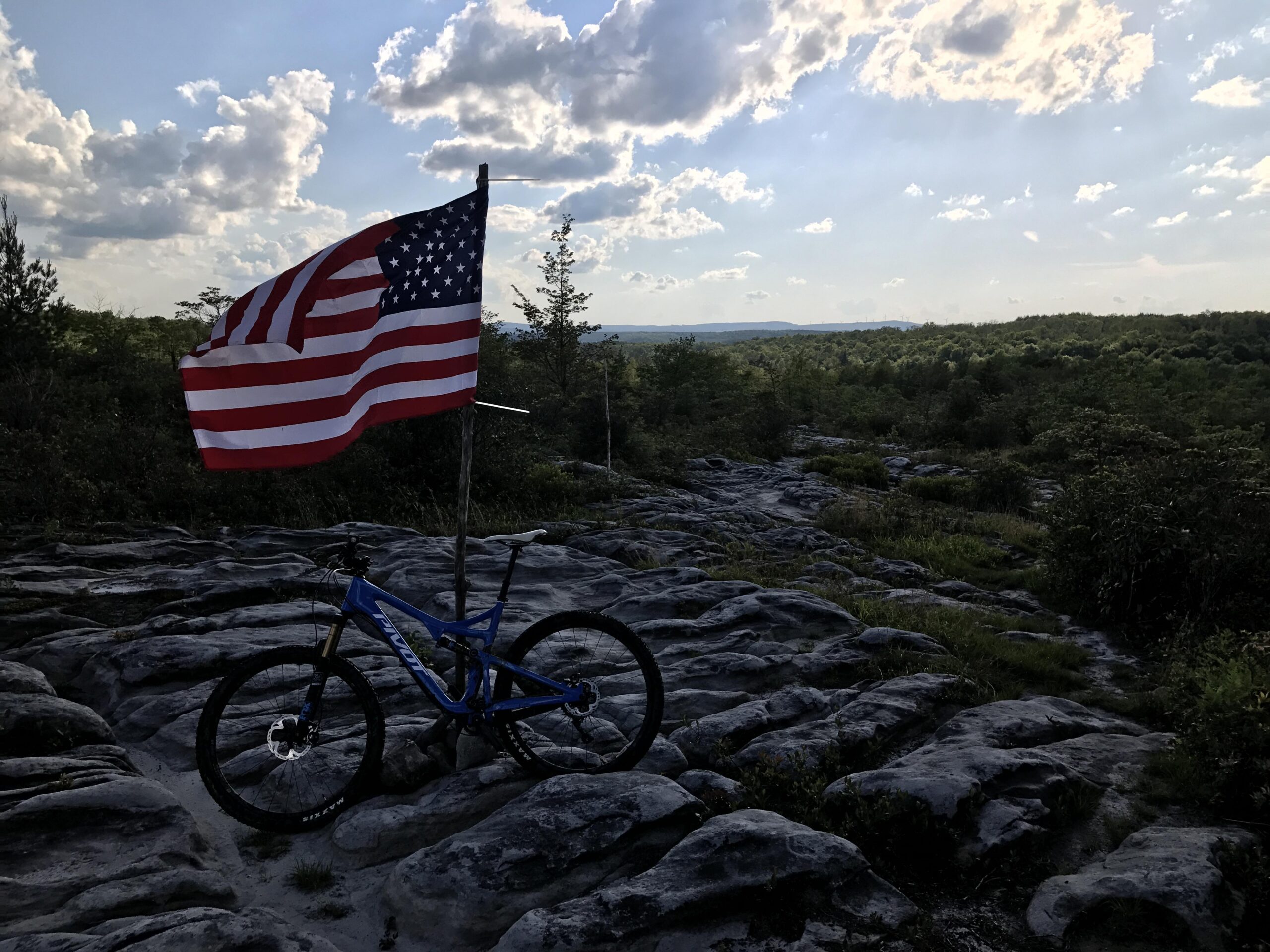 Pivot Trail 429: A mountain bike rests on a rocky terrain with a United States flag waving in the breeze. The scene features a backdrop of green trees and distant hills under a partly cloudy sky, capturing a moment of outdoor adventure and patriotism.