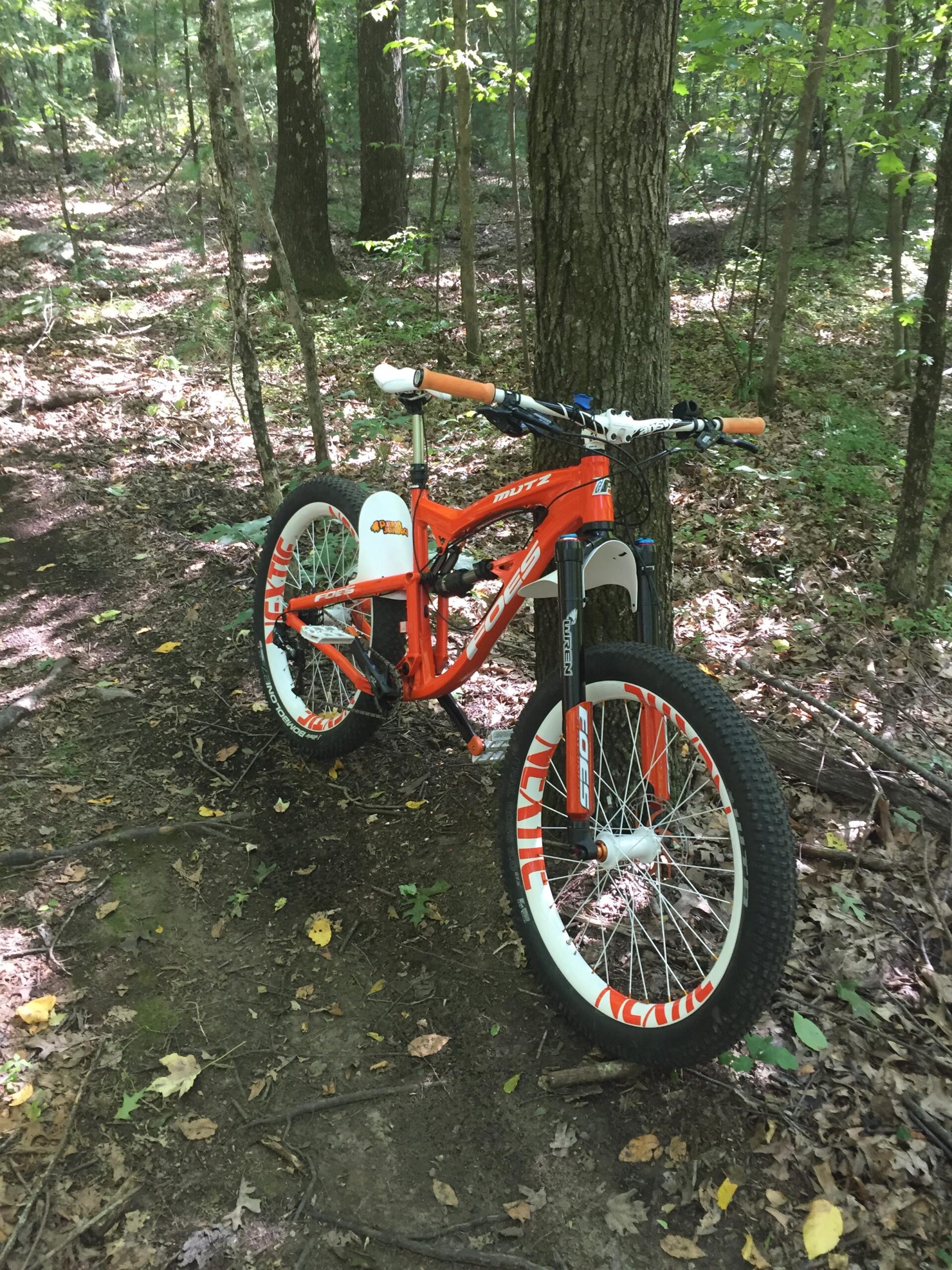 Foes Mutz: A bright orange mountain bike with distinctive white wheels is propped against a tree in a wooded area. The background features a mix of green foliage and brown earth, with sun filtering through the trees, creating a serene outdoor setting.