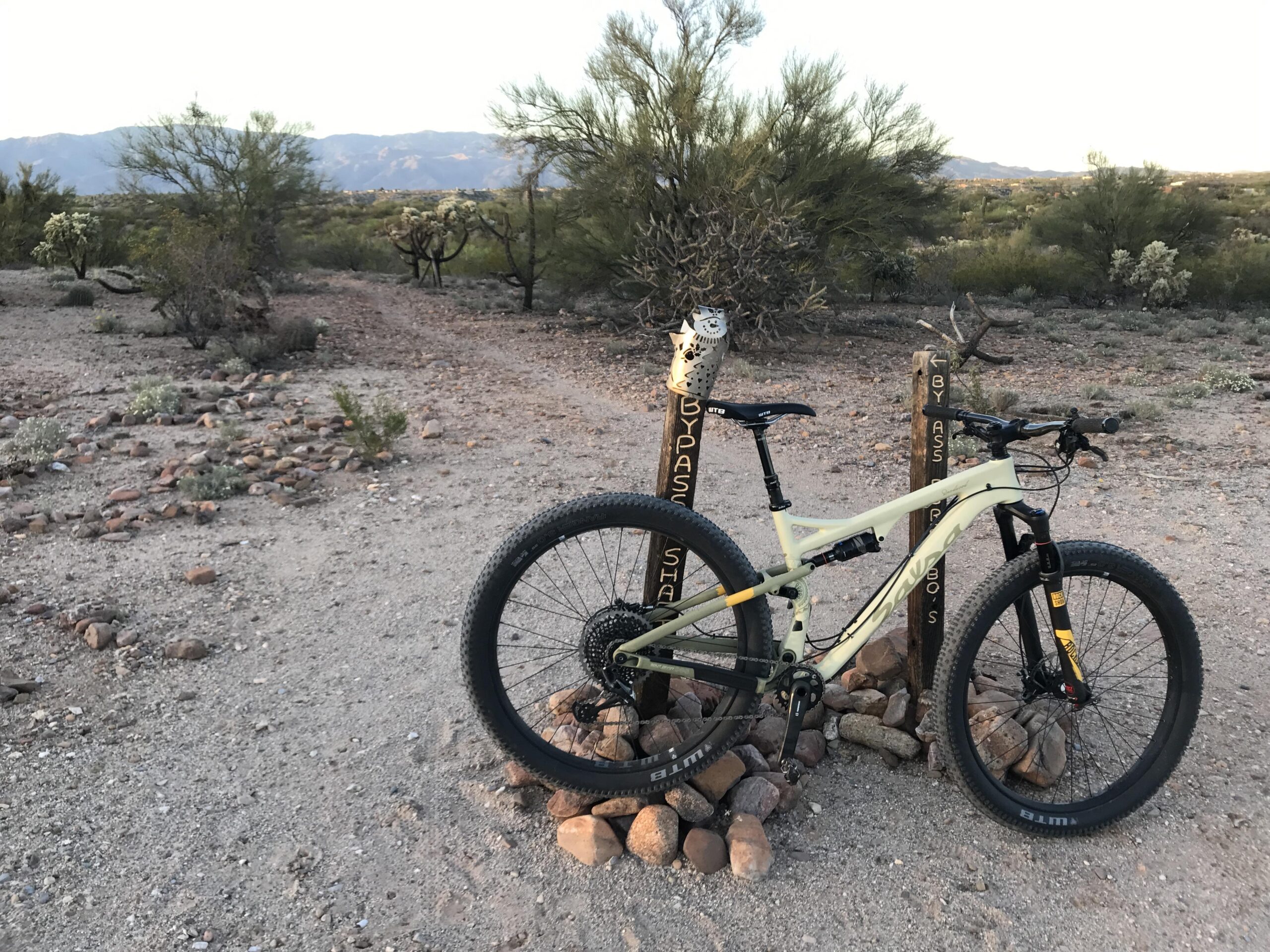 Salsa Deadwood Sus: A mountain bike parked on a rocky area in a desert landscape, with trail markers in the background. The scene features sparse vegetation and distant mountains under a clear sky, indicating an outdoor recreational location.