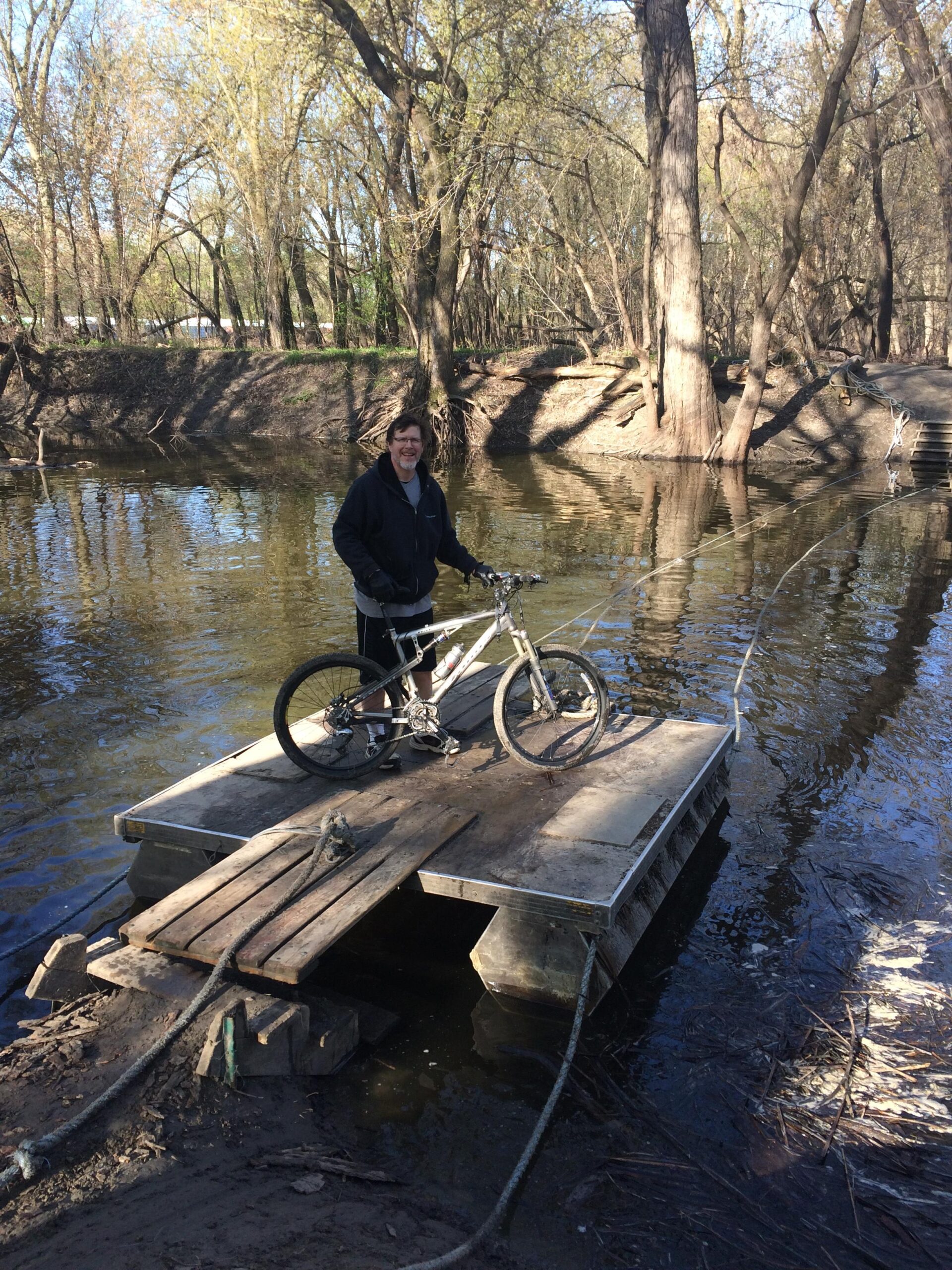 Jamis Dakar XC: A person stands on a small wooden platform over a calm river, holding a bicycle. They are smiling, wearing a black jacket and standing next to trees that line the riverbank, with sunlight filtering through the branches. The scene captures a peaceful outdoor setting in early spring.
