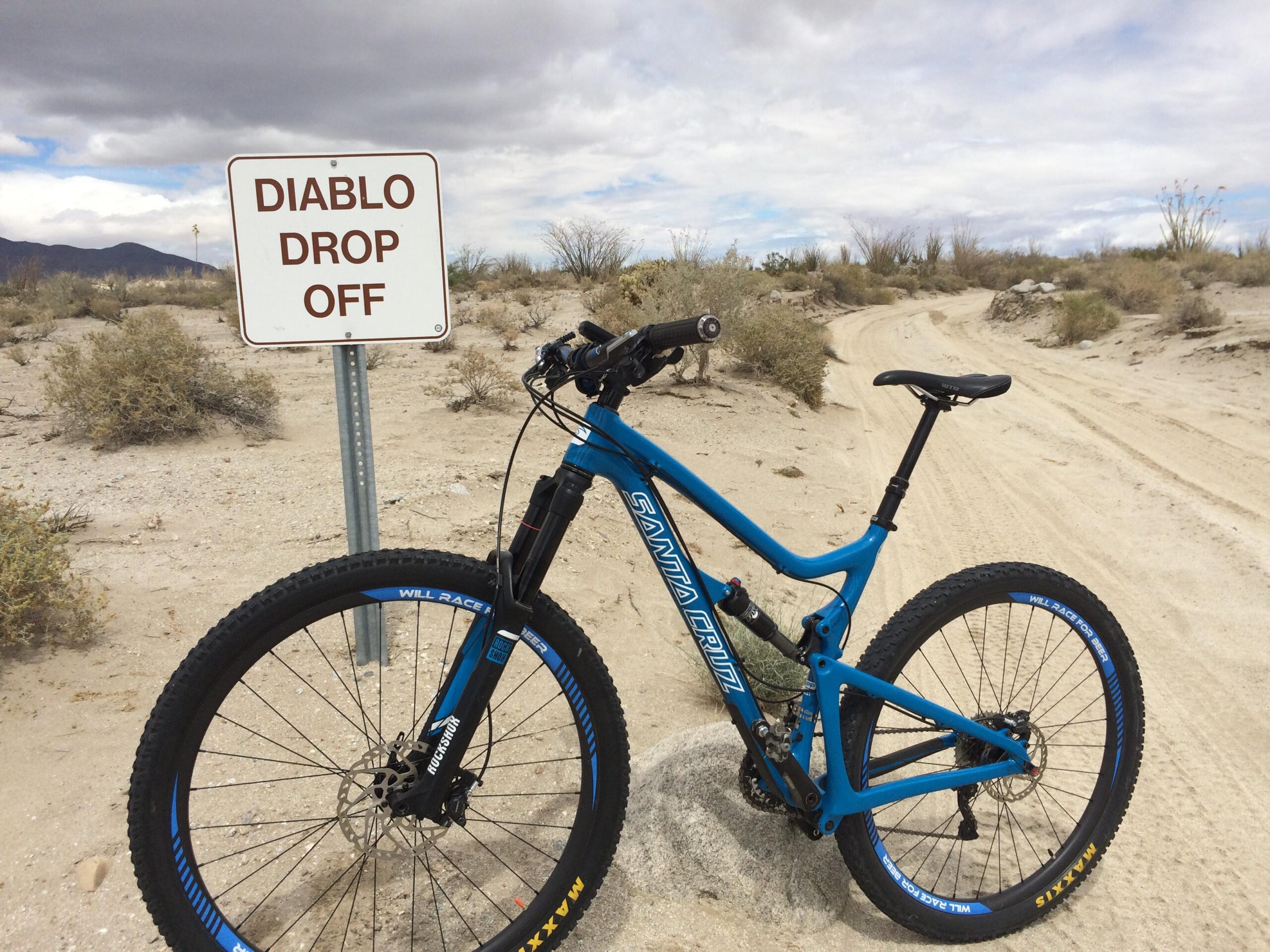 Santa Cruz Tallboy Carbon: A blue mountain bike parked next to a sign that reads "DIABLO DROP OFF," set against a sandy landscape with sparse vegetation and a cloudy sky. A dirt trail winds in the background.