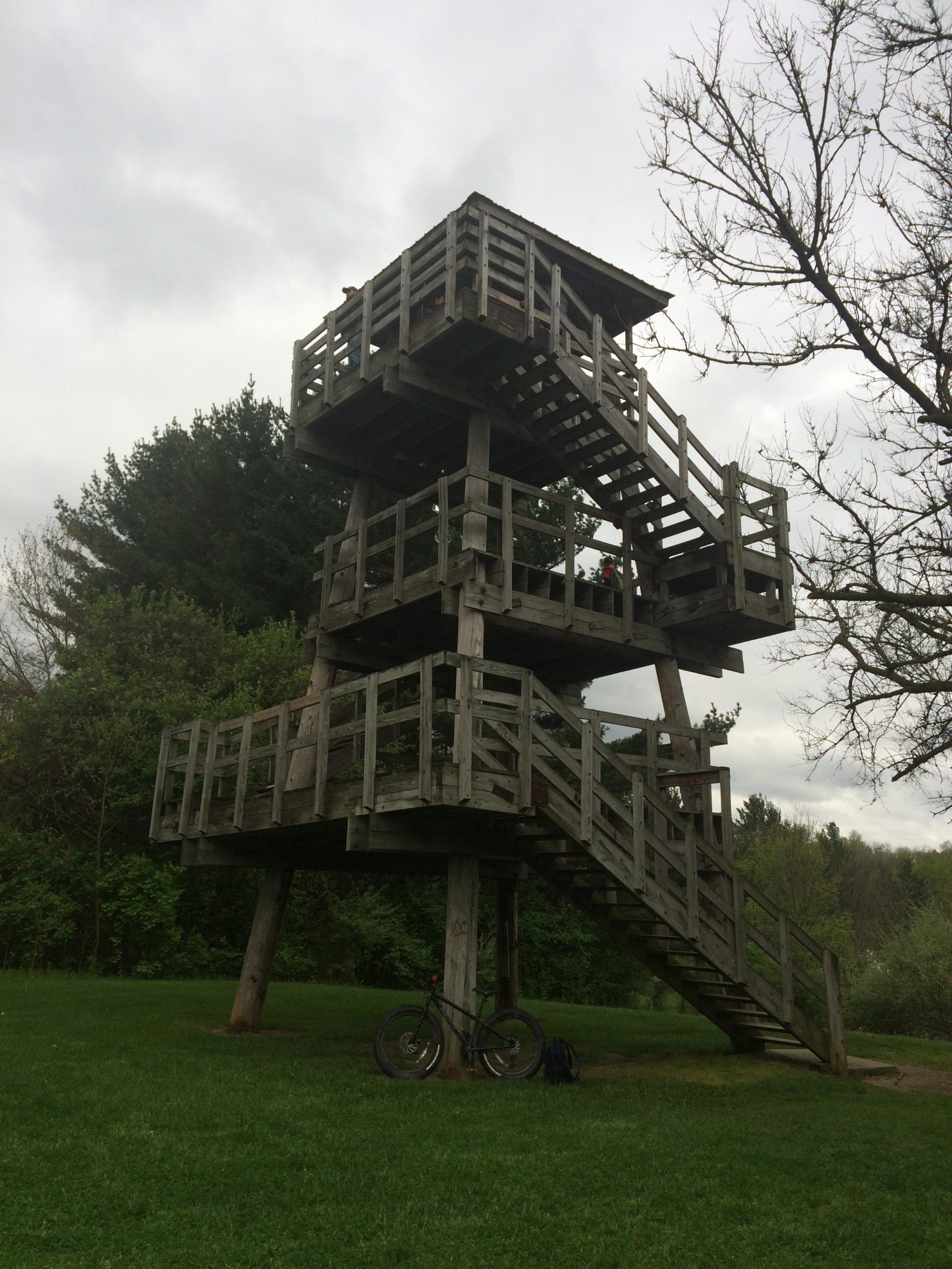A wooden observation tower with multiple levels, surrounded by green grass and trees, under a cloudy sky. A bicycle leans against the tower, and a backpack is positioned nearby. Bonneyville Mill mountain bike trail.