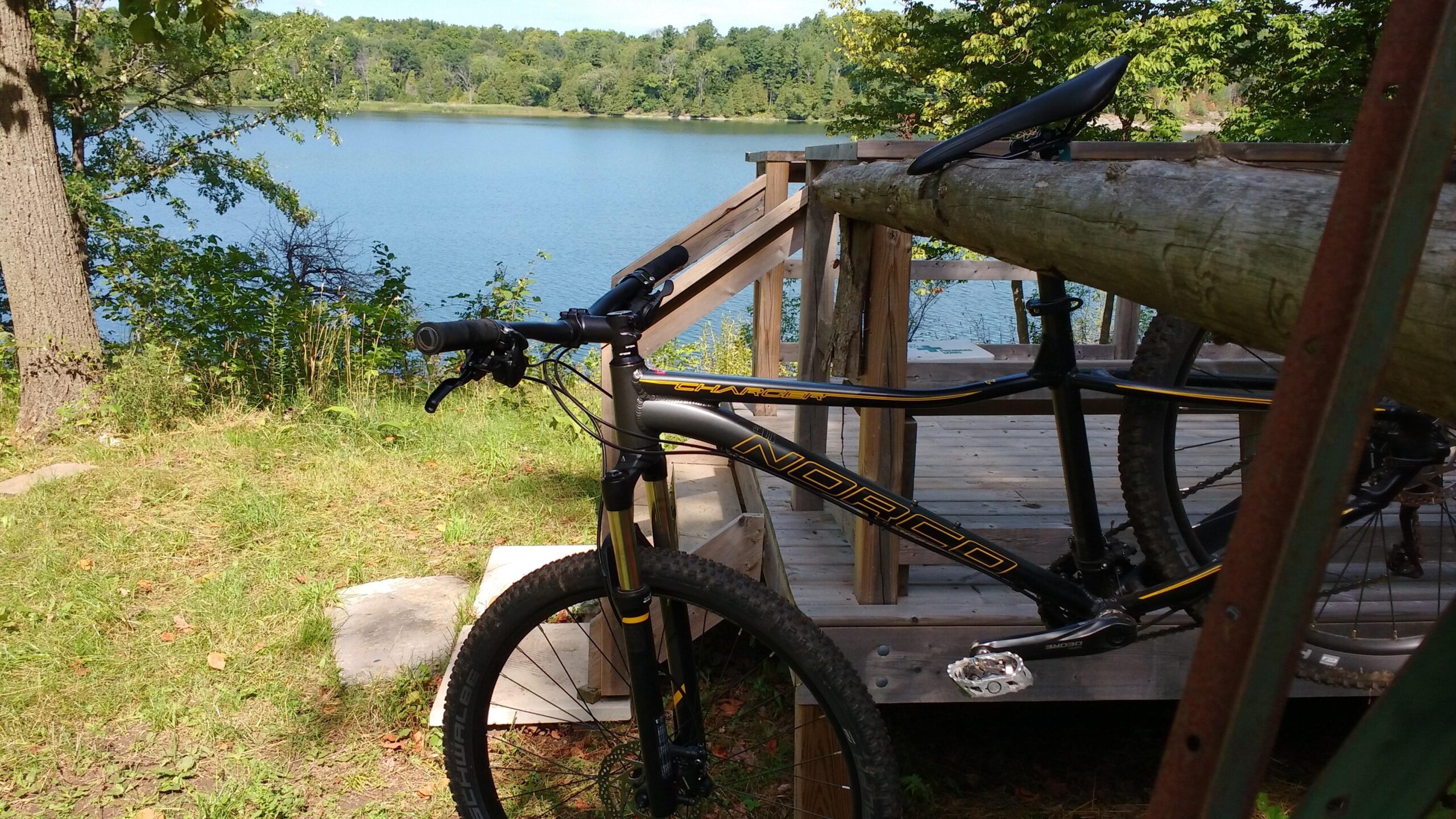 Norco Charger 7.2: A mountain bike rests on a wooden deck overlooking a tranquil lake surrounded by trees and greenery on a sunny day.