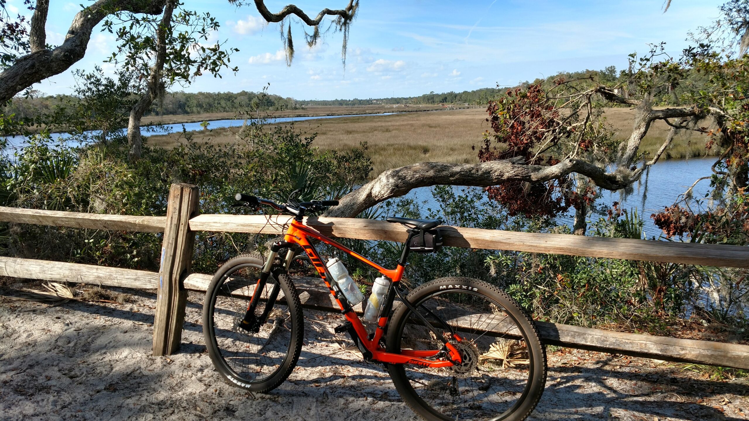 Giant Giant Fathom 2 29er: A mountain bike with orange and black accents is parked against a wooden fence, overlooking a scenic view of a marshland and waterway surrounded by trees and vegetation. The sky is clear with a few clouds, and the landscape features tall grasses and shrubs, creating a serene outdoor atmosphere.