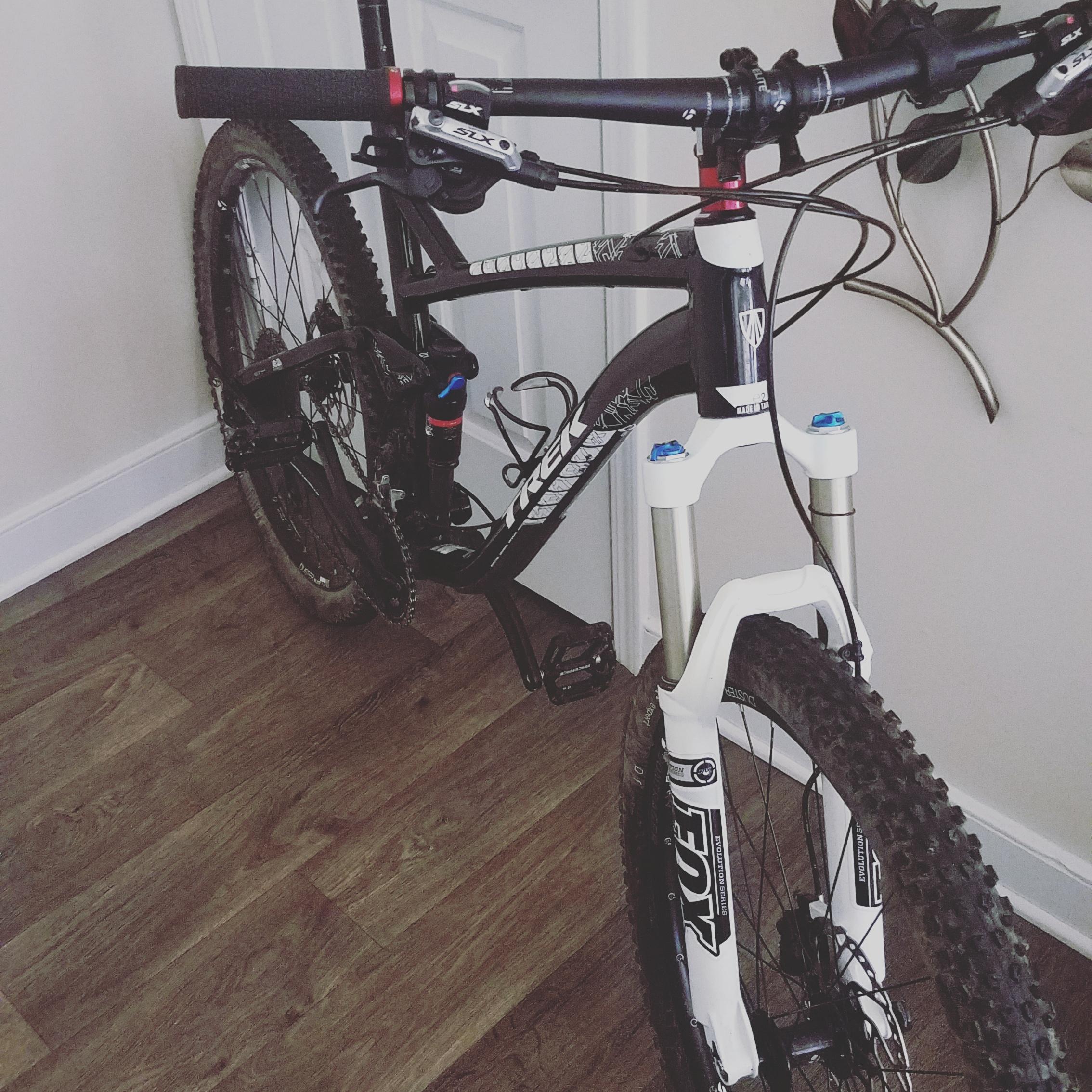 Trek Fuel EX 8: A black and white mountain bike leaning against a wall, featuring a front suspension fork, handlebars, and visible pedals. The bike is resting on a wooden floor, with dirt on the tires indicating recent use.