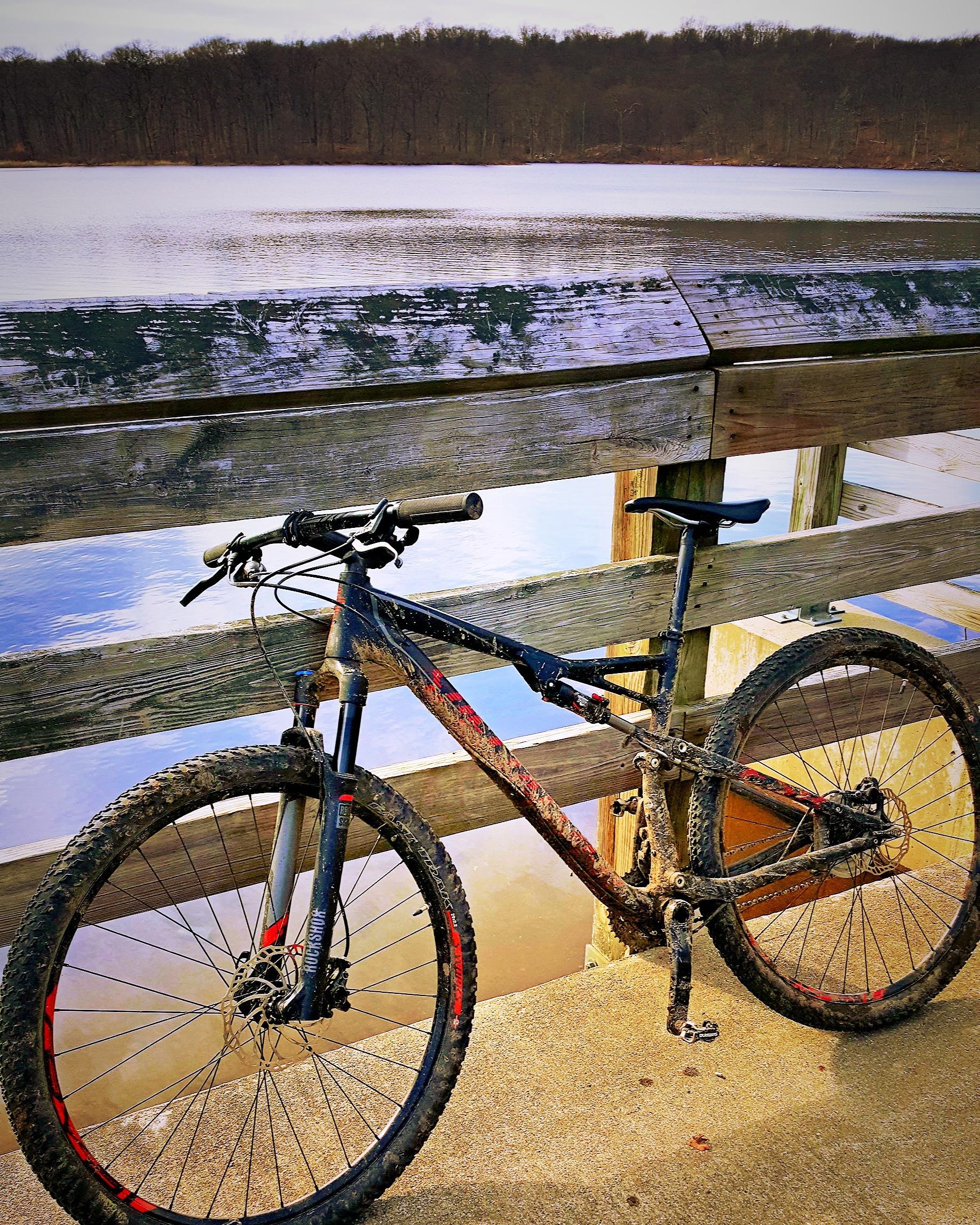 A mountain bike with dirt on its frame and tires is positioned on a wooden dock by a lake. The calm water reflects the surrounding trees, and the scene is illuminated by soft, natural light. Allamuchy Mt. State Park: Deer Park mountain bike trail.