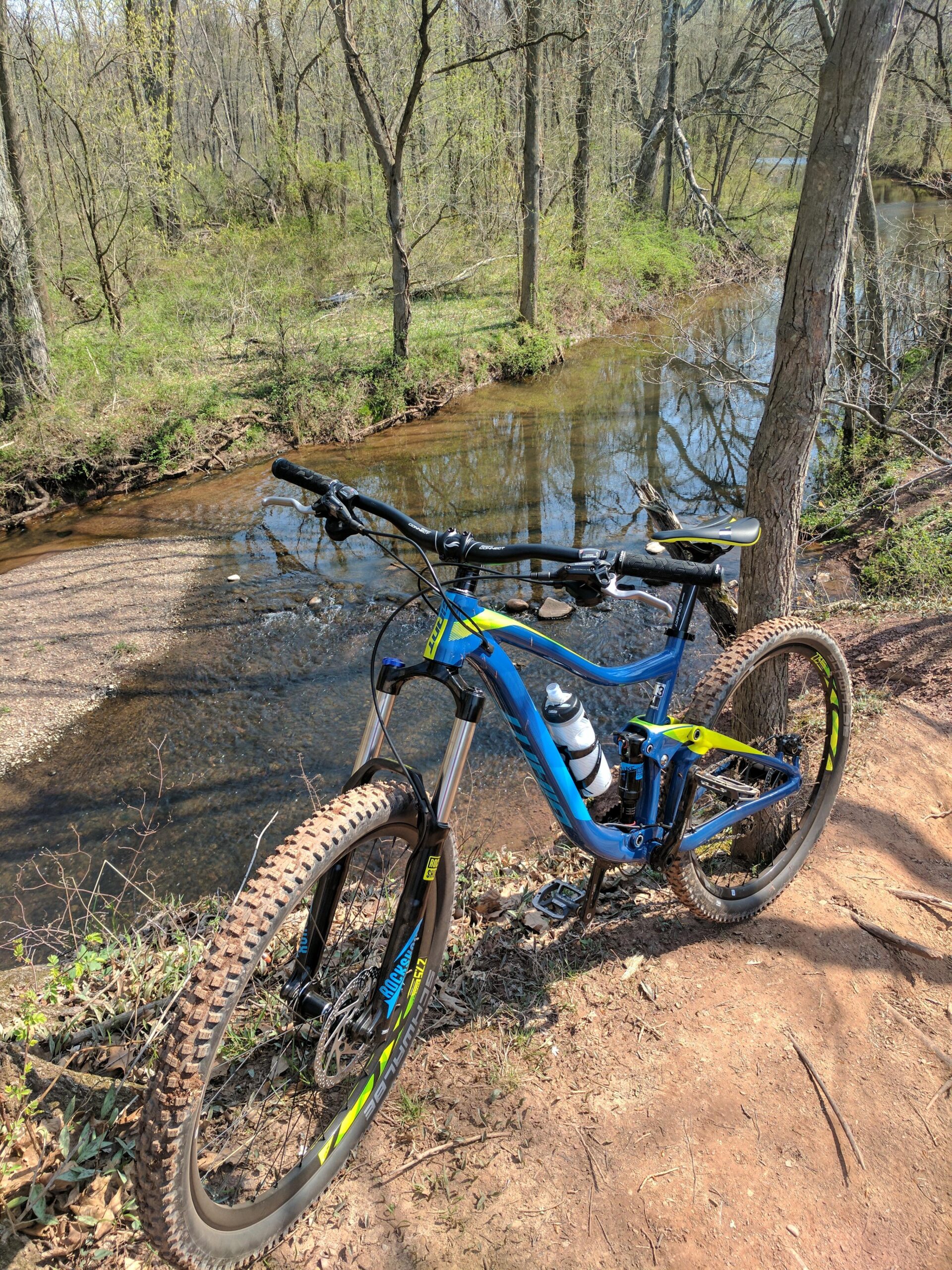 Giant Trance: A mountain bike resting beside a calm, reflective stream in a wooded area. The bike features a blue and yellow design, with thick, treaded tires positioned on a dirt path surrounded by greenery. Tall trees with budding leaves frame the scene, indicating a springtime atmosphere.