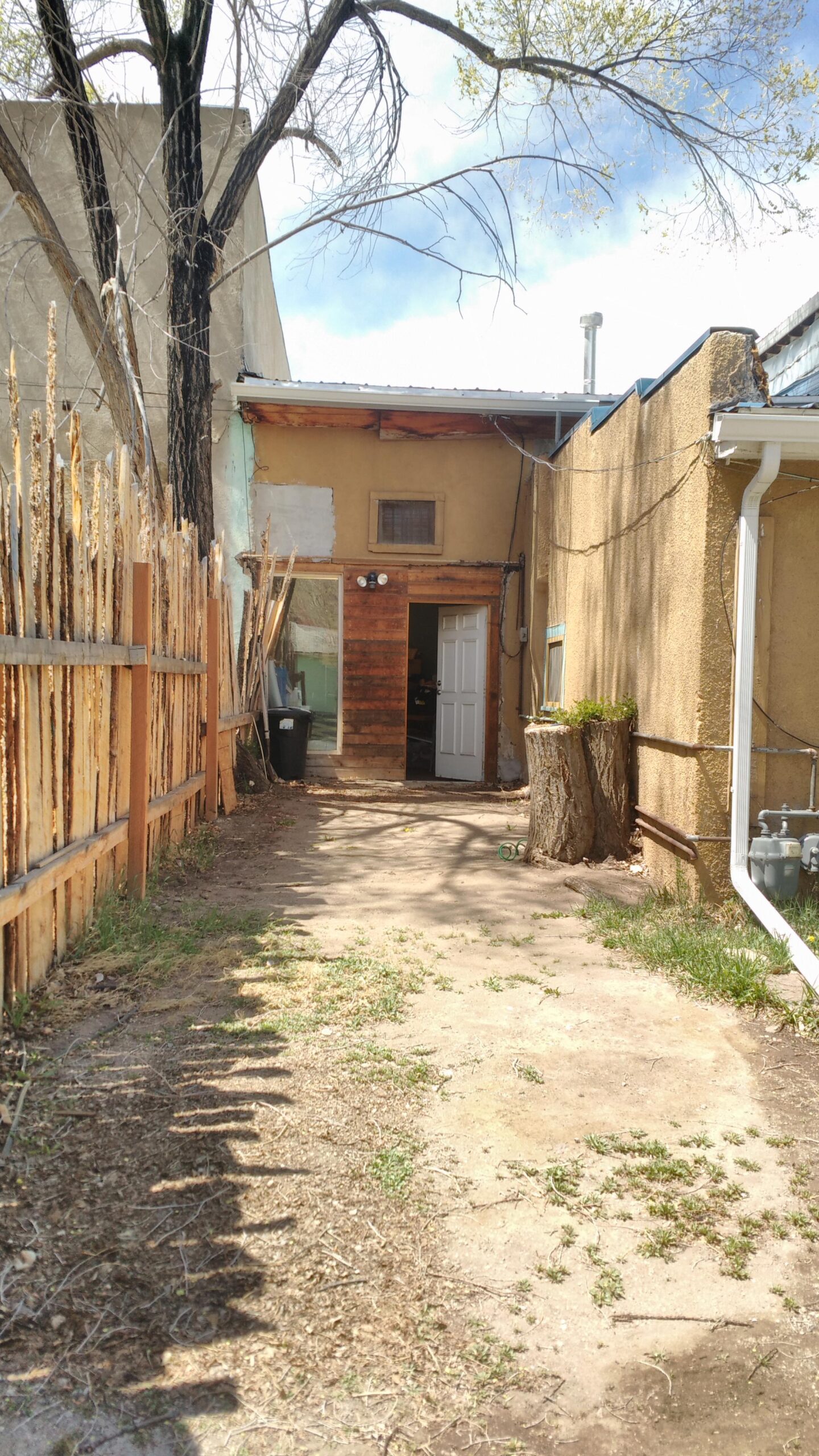 A narrow dirt pathway leads towards the entrance of a rustic building, framed by a wooden fence on the left. A tall, barren tree is located on the left side, and the building features a wooden door and a small window. To the right, there