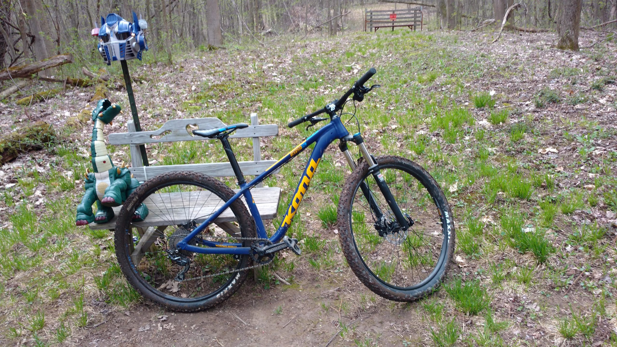 A blue mountain bike is parked next to a wooden bench in a forested area, with green grass and fallen leaves on the ground. On the bench, there is a colorful inflatable dinosaur toy and an Optimus Prime figure. A rustic gate is visible in the background, indicating a trail or pathway. Great Bear mountain bike trail.