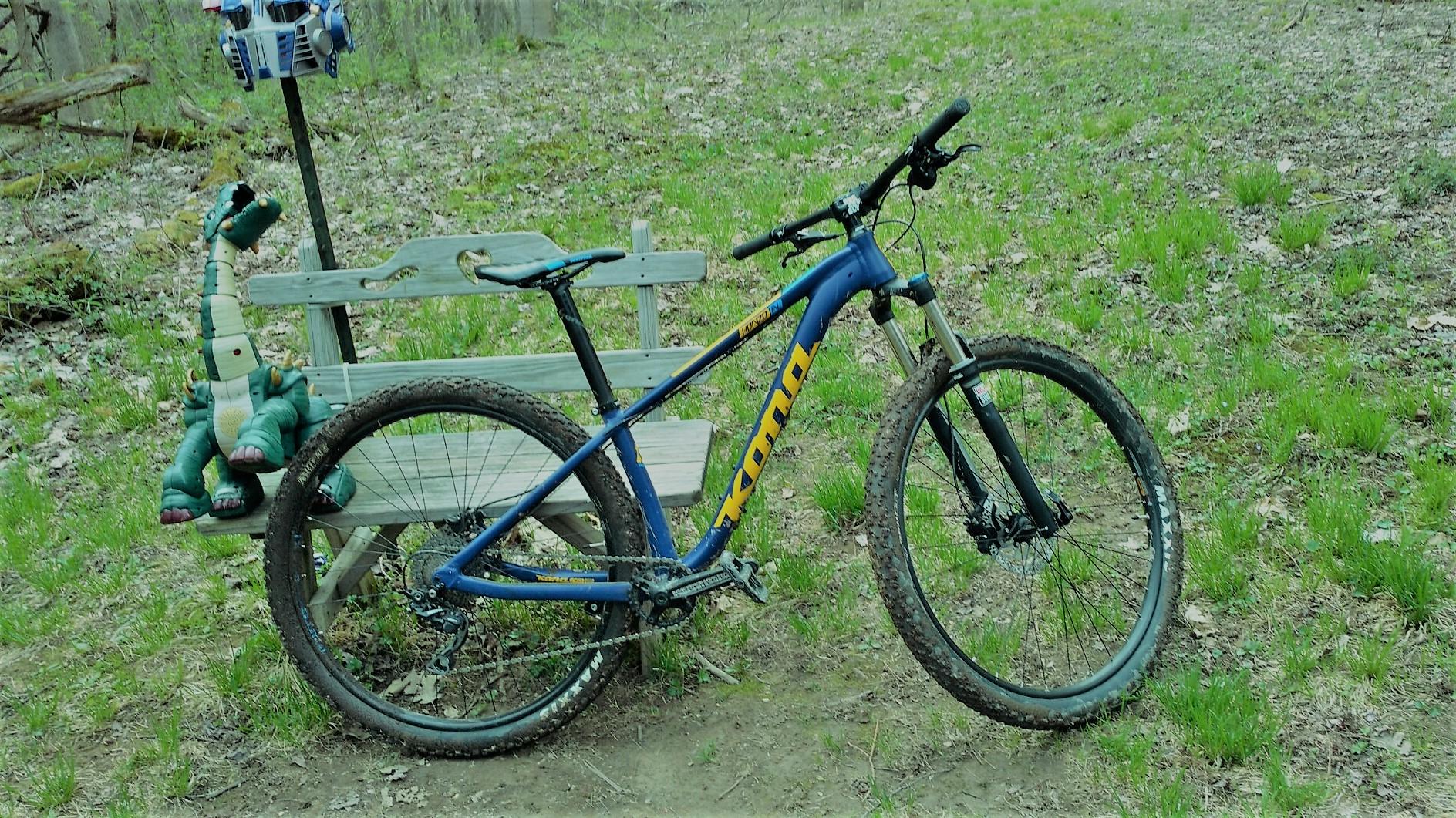 A blue mountain bike leaning against a wooden bench in a grassy area, accompanied by a plush dinosaur toy sitting beside it. In the background, a tree line is visible, suggesting a natural outdoor setting. Great Bear mountain bike trail.