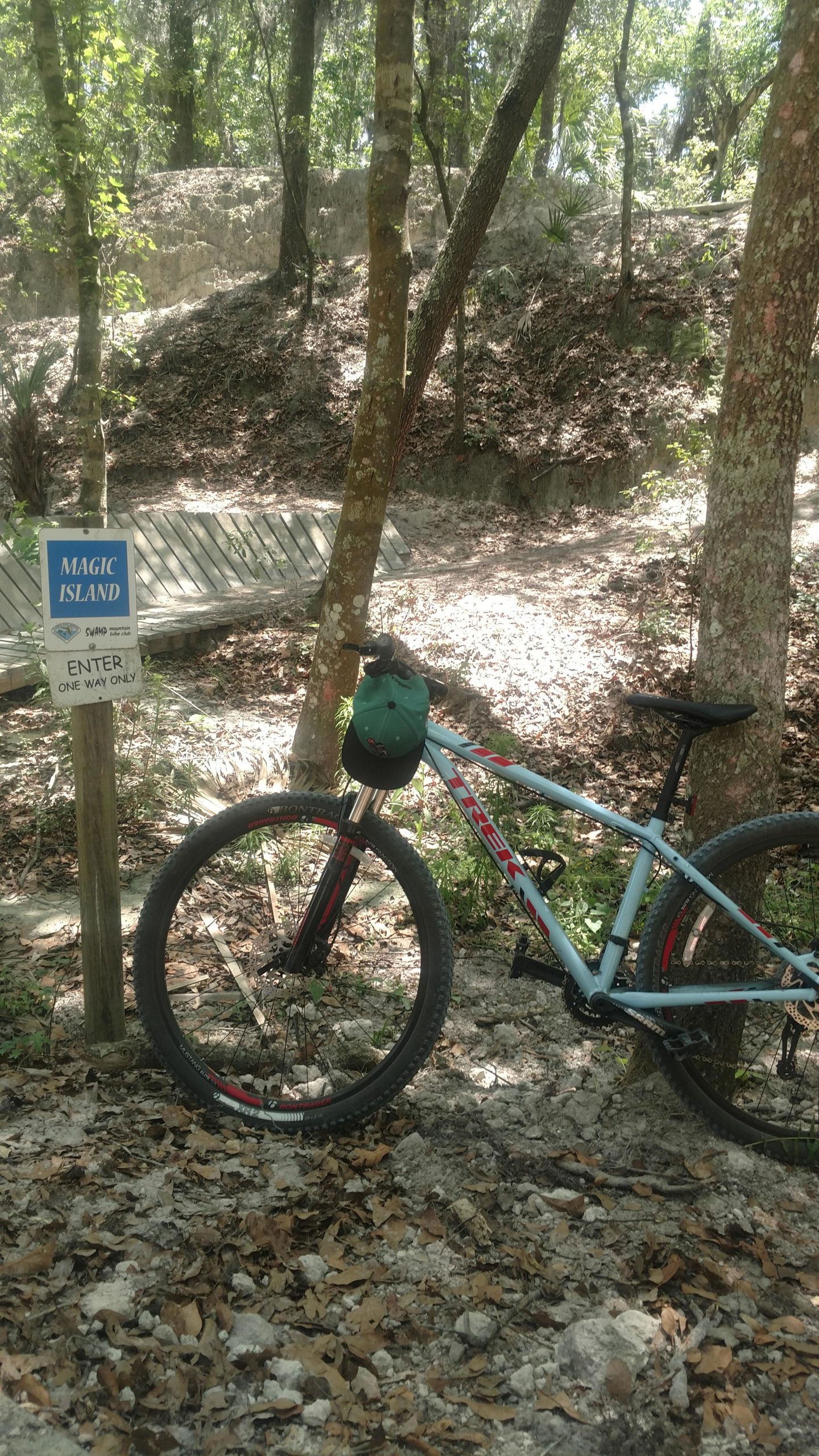 A mountain bike resting against a tree near a sign that reads "Magic Island, Enter One Way Only." The surrounding area is a wooded path with leaves and rocky terrain, indicating a natural outdoor setting. Alafia River State Park mountain bike trail.