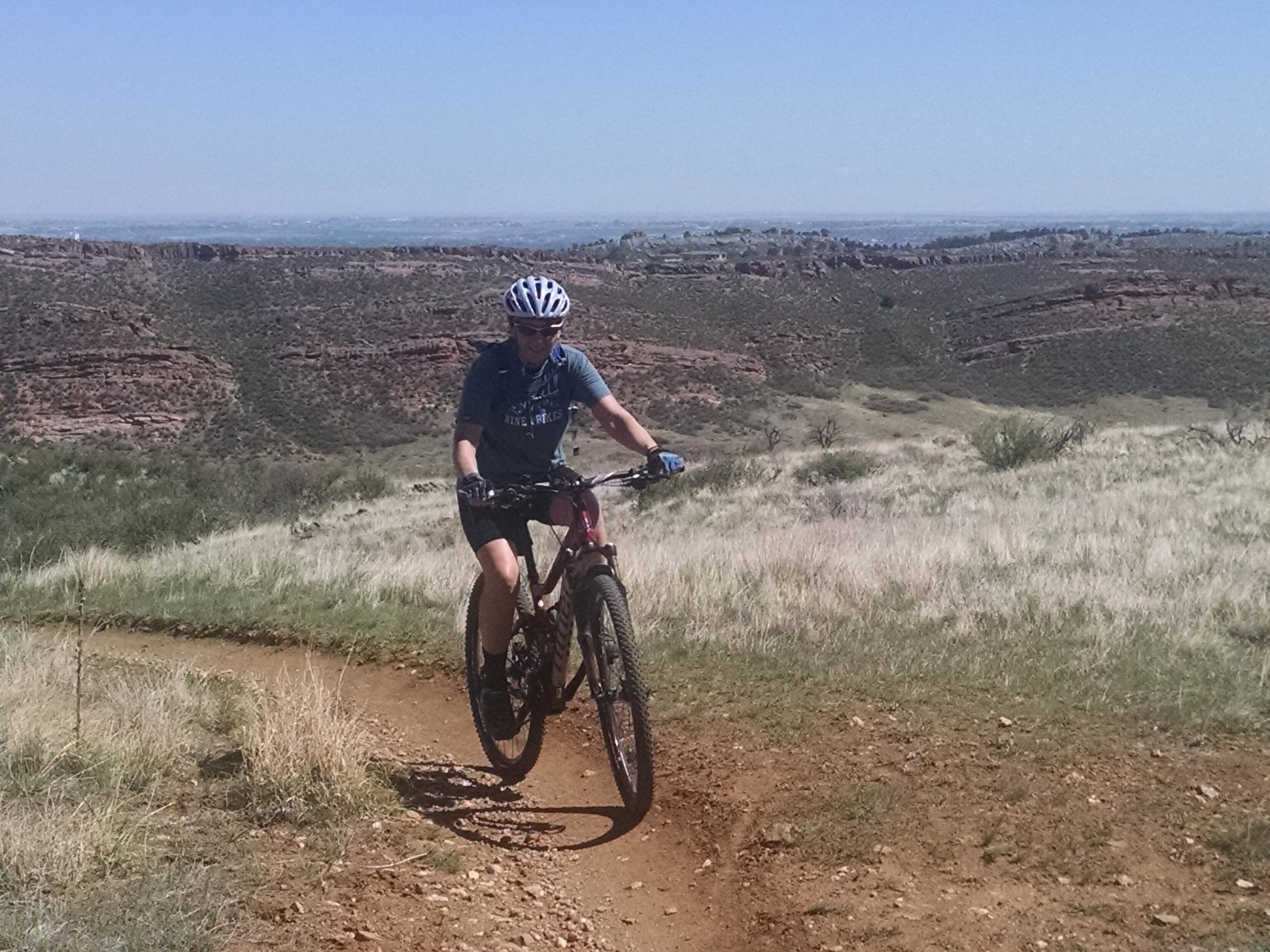 A person riding a mountain bike on a dirt trail surrounded by grassy fields and rocky hills under a clear blue sky. Blue Sky mountain bike trail.