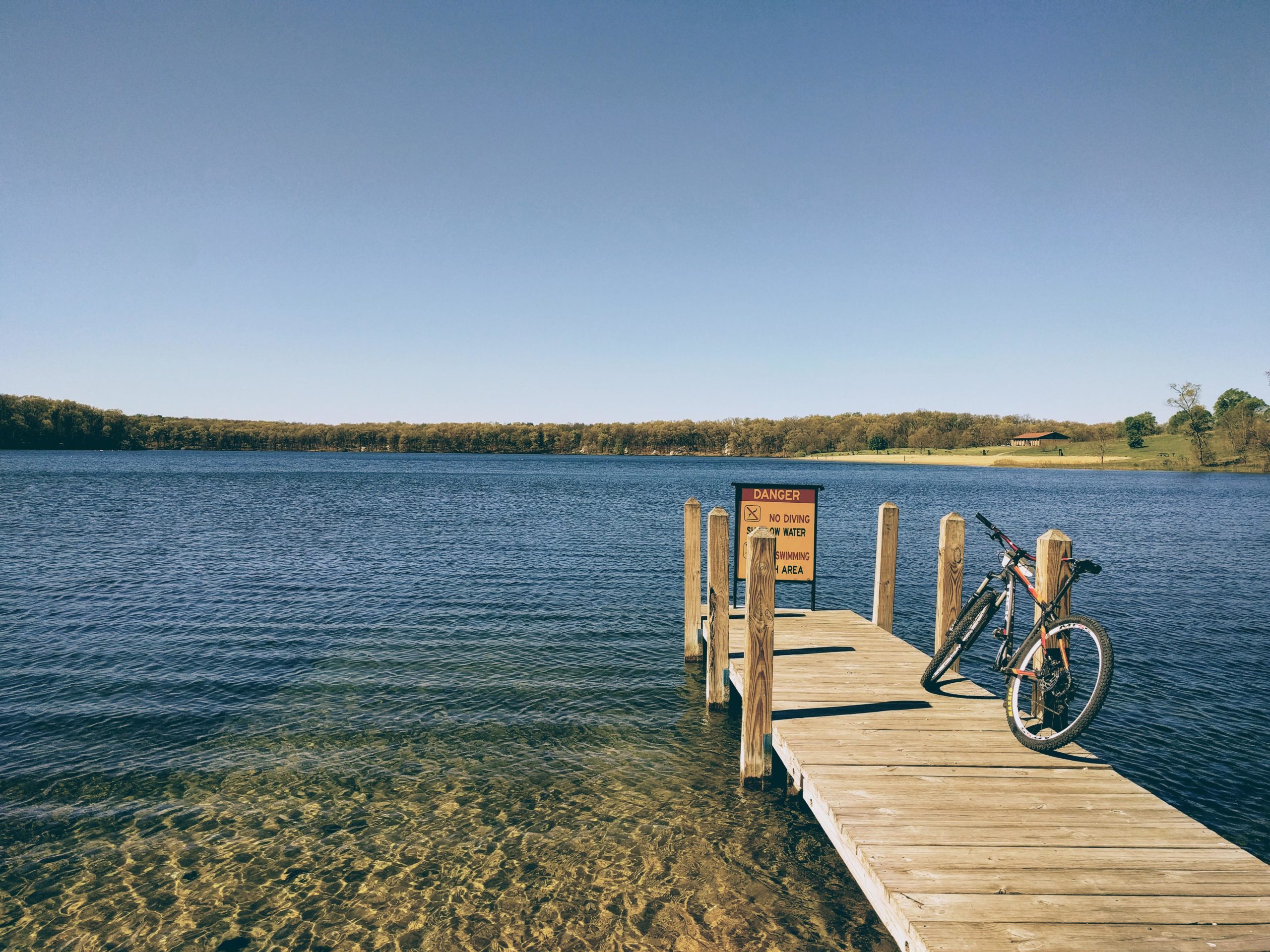 A wooden dock extends into a calm lake, with a mountain bike resting against the side. A warning sign on the dock indicates danger, with notes about no diving or swimming. The scene features clear blue skies and a lush green forest in the background, reflecting a peaceful outdoor setting. Fort Custer Recreation Area mountain bike trail.