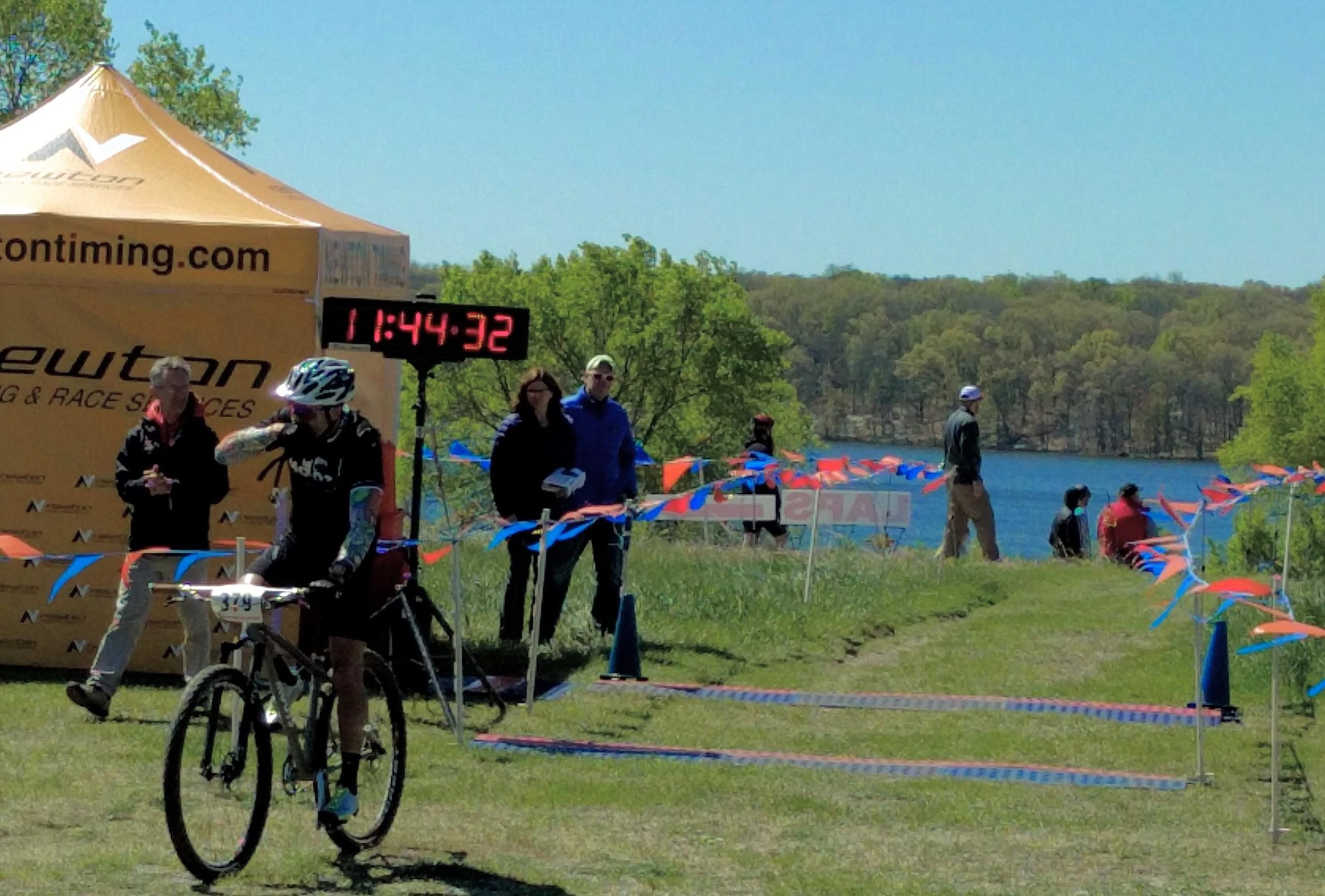 A cyclist crossing the finish line at a race, with a digital timer displaying a time of 11:44:32. In the background, spectators stand near a tent, and there is a scenic view of a lake and trees. Colorful flags and cones mark the race course. Fort Custer Recreation Area mountain bike trail.