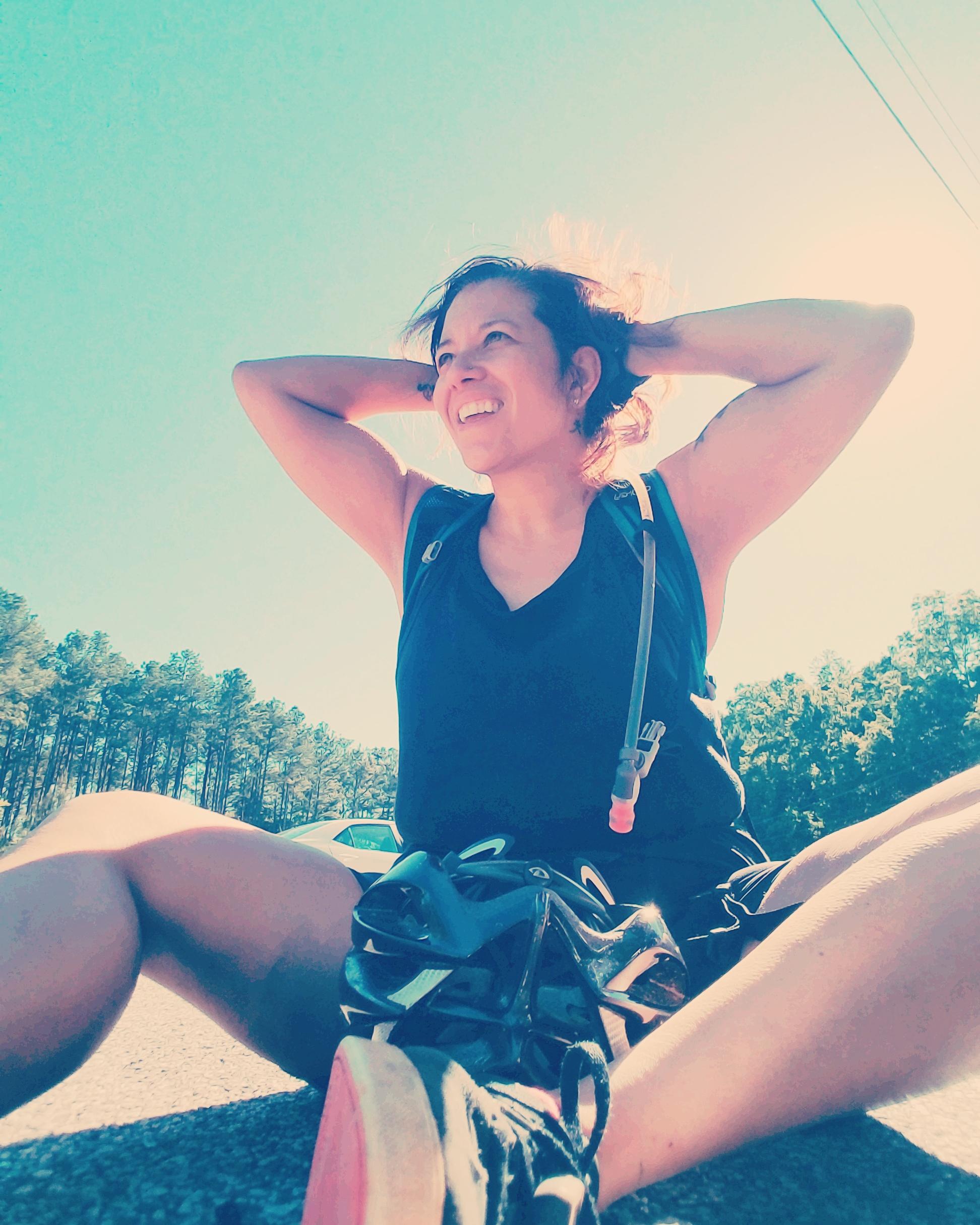 A person sitting on the ground outdoors, smiling and enjoying the sunny weather. They are wearing a black tank top and are surrounded by pine trees in the background. A backpack is partially visible, and the person has one hand behind their head. The scene conveys a sense of relaxation and joy. Fort Yargo State Park mountain bike trail.