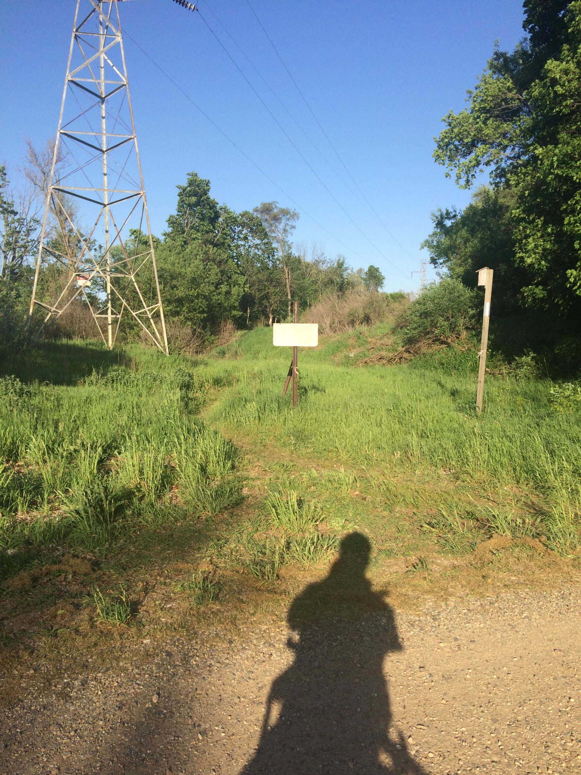 A dirt path leading through an overgrown area, with a large power line tower on the left and two signs alongside the path. The sky is clear and blue, and a person's shadow is visible in the foreground on the gravel road. The Dump mountain bike trail.