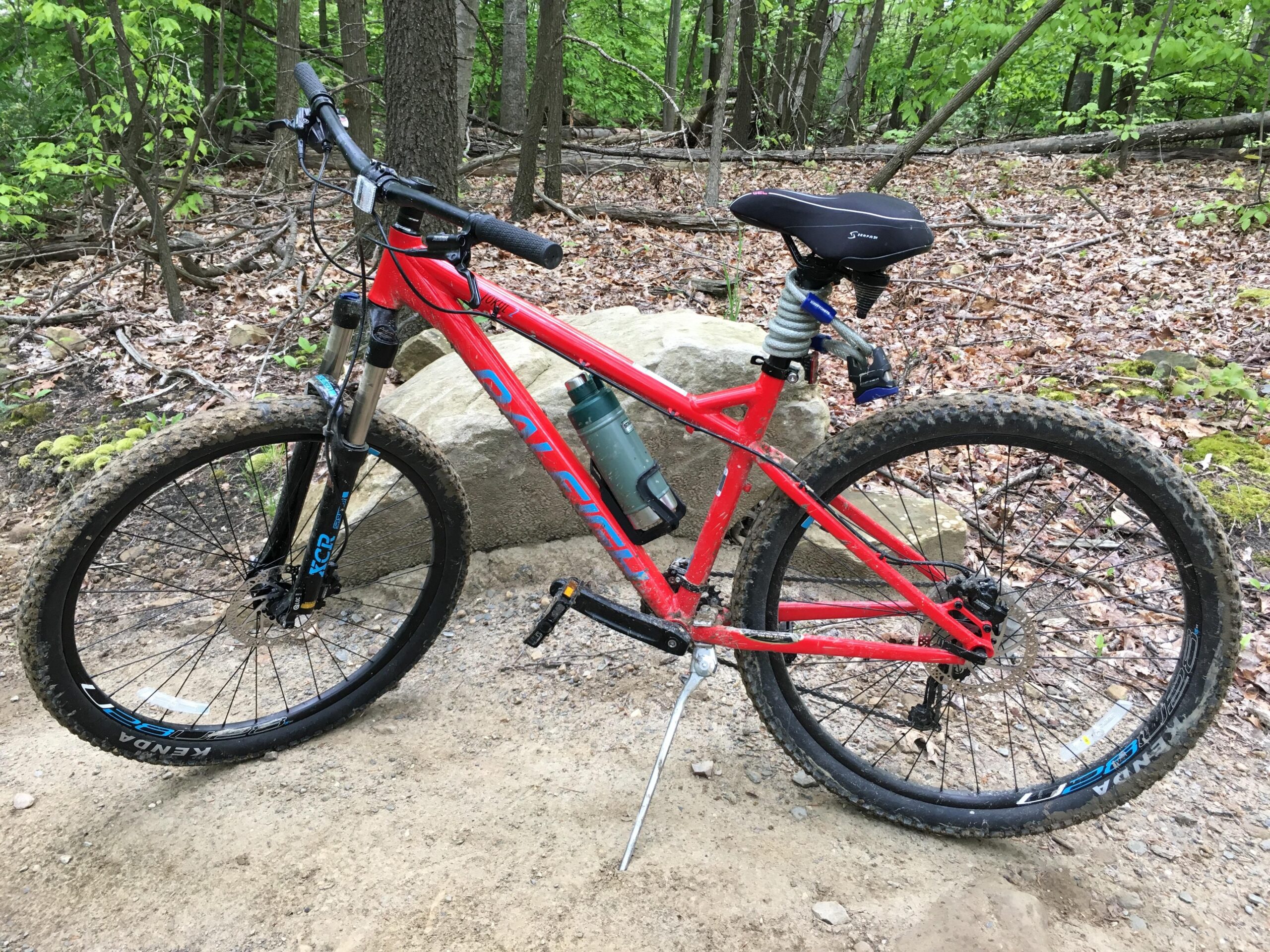 Raleigh Tokul 2: A red mountain bike with a front suspension fork, set against a backdrop of green foliage and a dirt trail. The bike has visible mud on the tires, indicating recent use on rugged terrain, and a water bottle attached to the frame. A large rock is partially in view beside the bike.