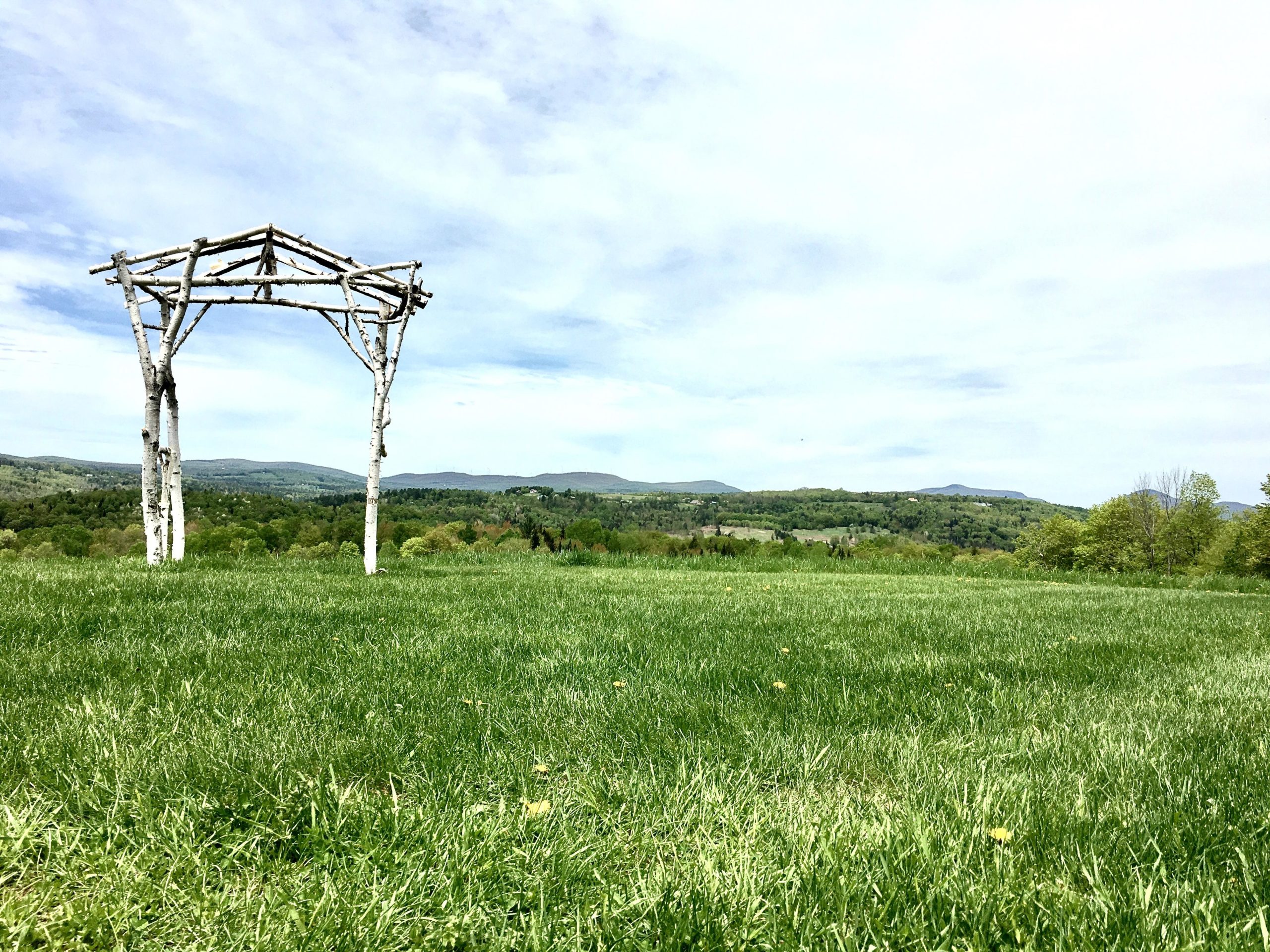 A rustic wooden arch stands in a lush green field, framed by rolling hills and a clear blue sky. The landscape showcases vibrant trees and gentle slopes in the background, creating a serene natural setting. Kingdom Trails mountain bike trail.